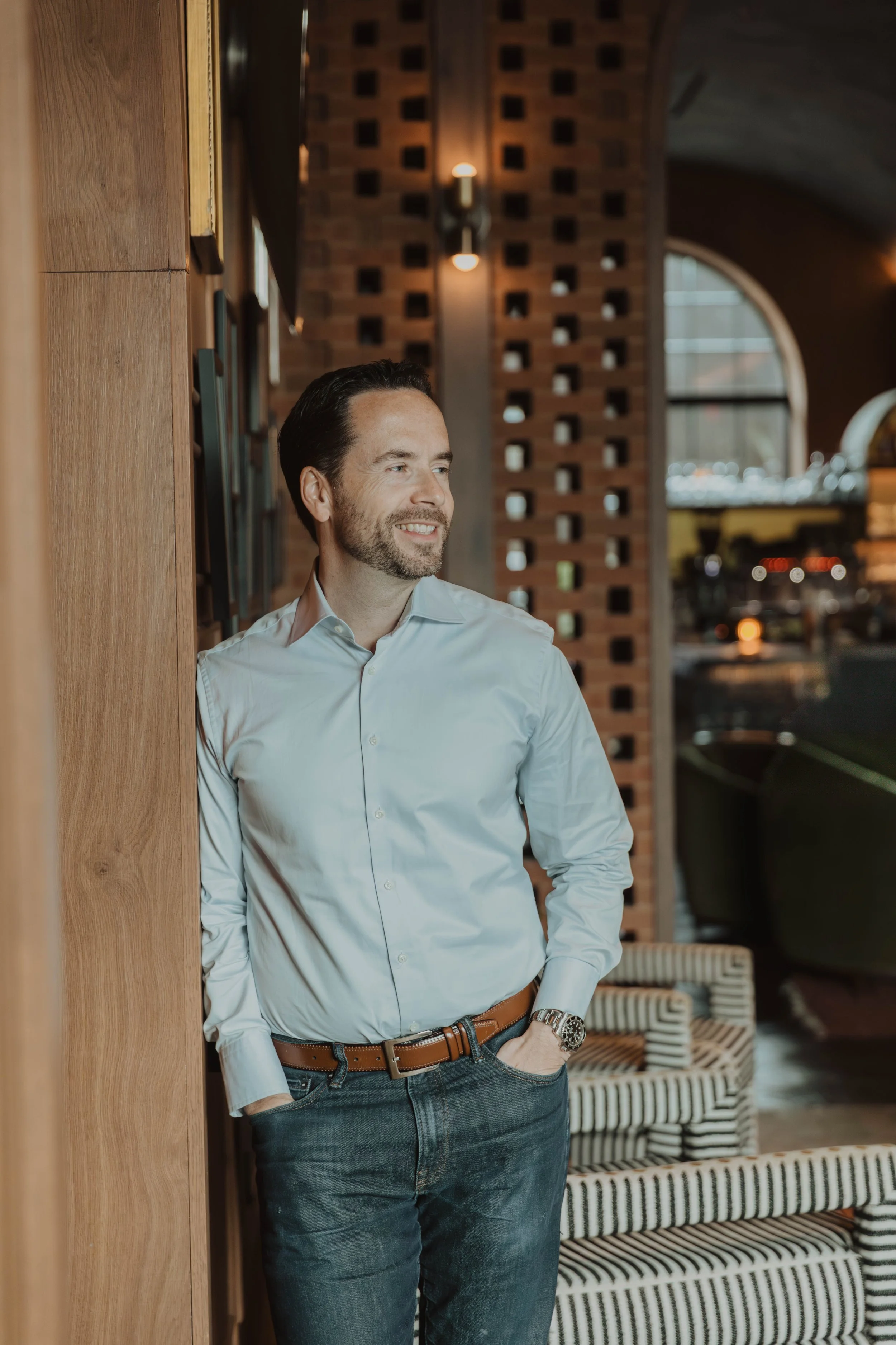 A man in a white shirt and jeans leaning against a wooden wall inside a modern, stylish restaurant or lounge, smiling and looking to the side.