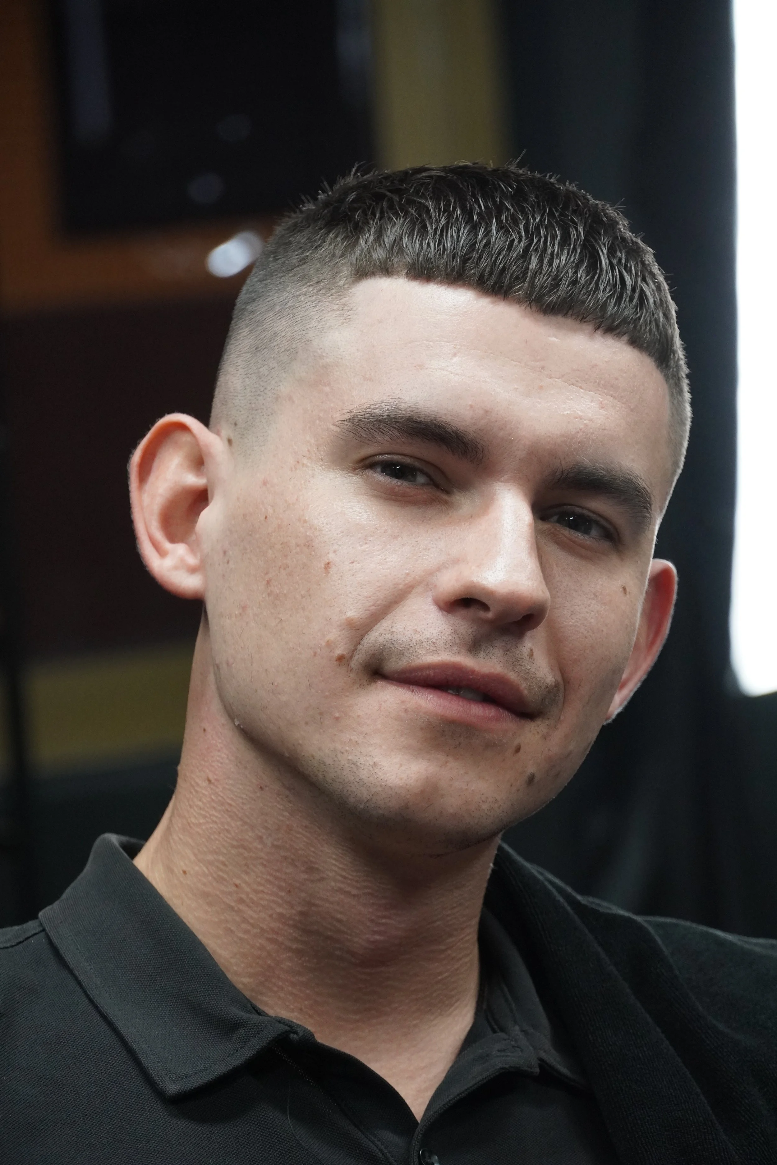 Close-up of a young man with short dark hair and light skin, wearing a black collared shirt, smiling slightly, indoors with a dark background.
