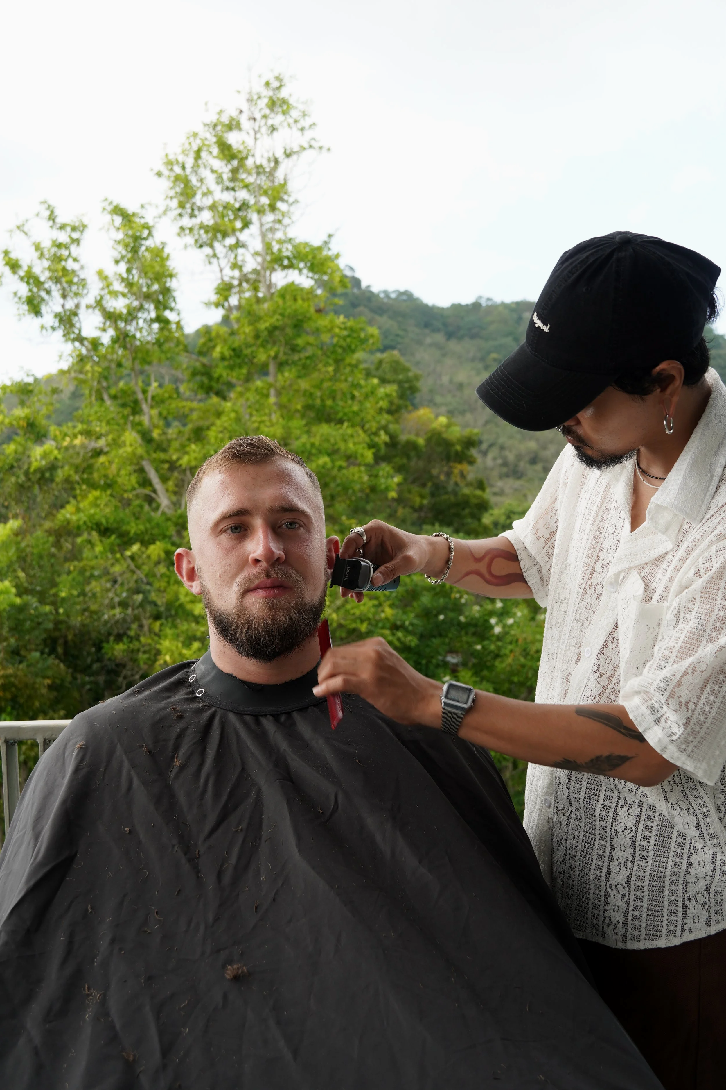 A man getting a haircut outdoors, with a scenic green mountain background.