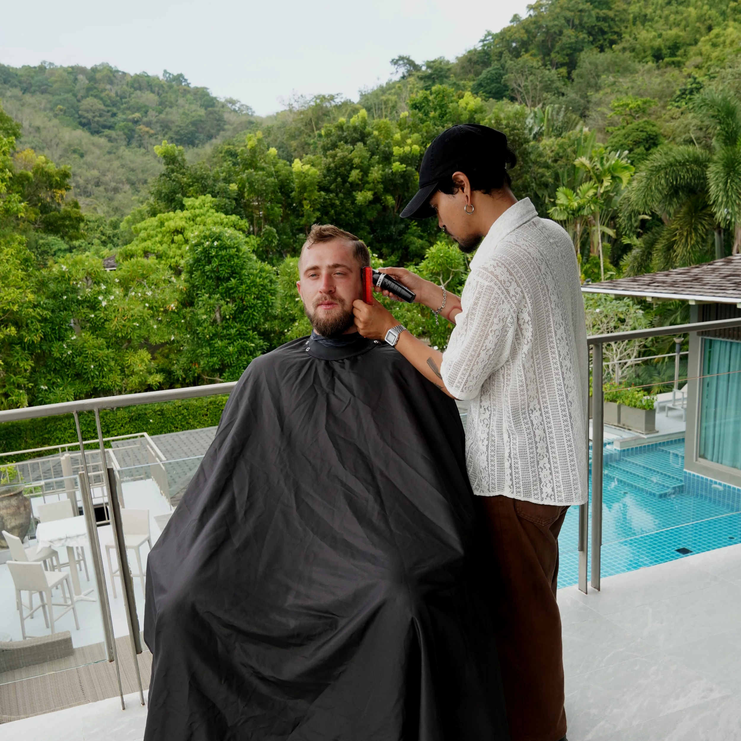 A man getting a haircut outdoors on a balcony with a scenic view of lush green trees and hills, while a barber trims his beard with electric clippers.