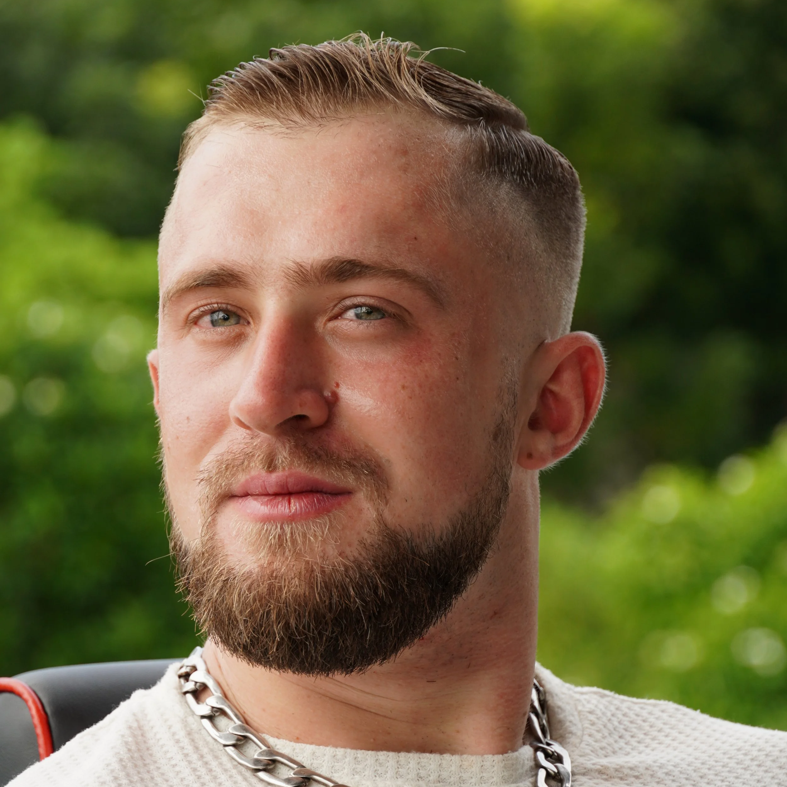 Close-up of a young man with a beard, blond hair, and piercing blue eyes, wearing a silver chain necklace, with a blurred green foliage background.