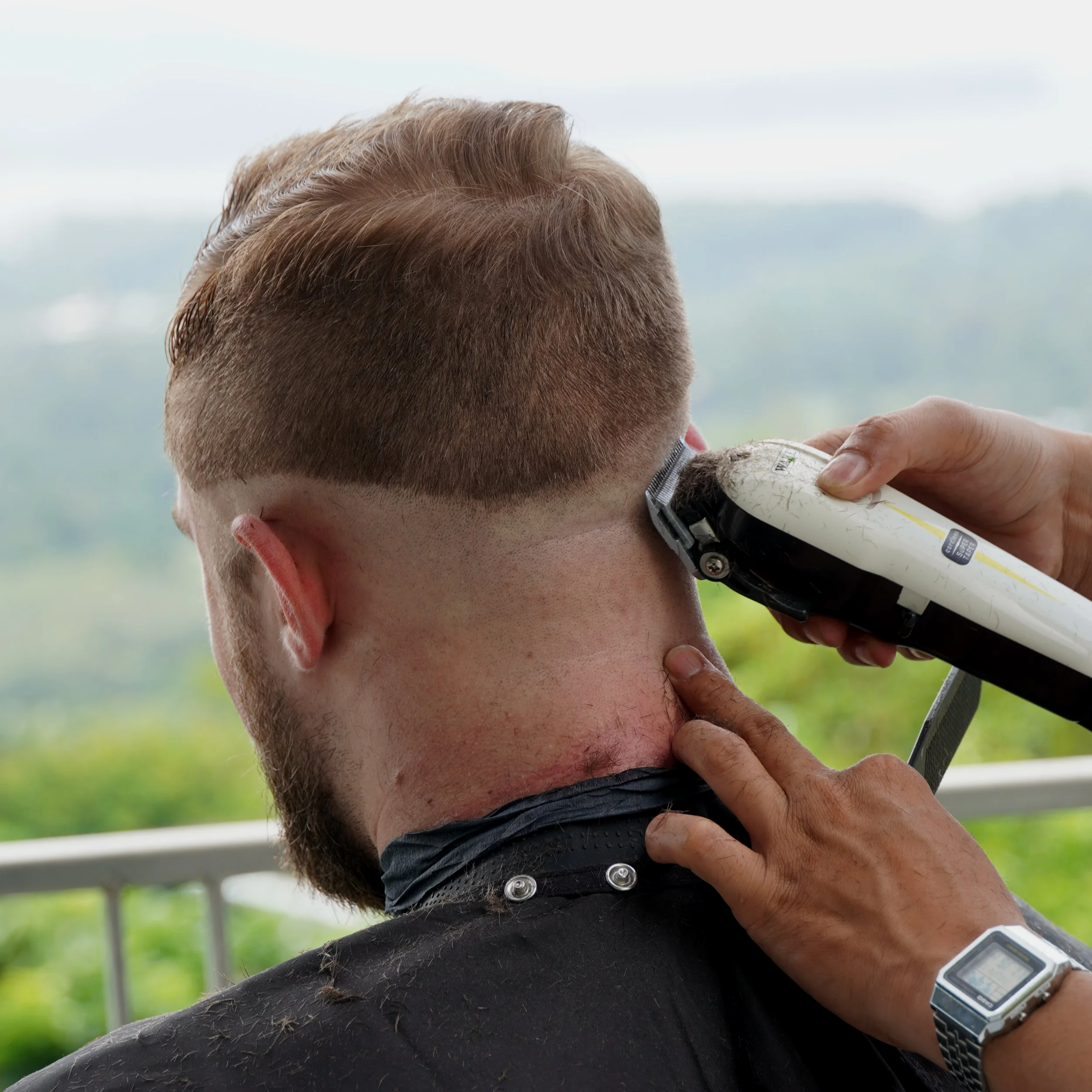 A man getting a haircut outdoors, using electric clippers to cut hair on the back of his head.