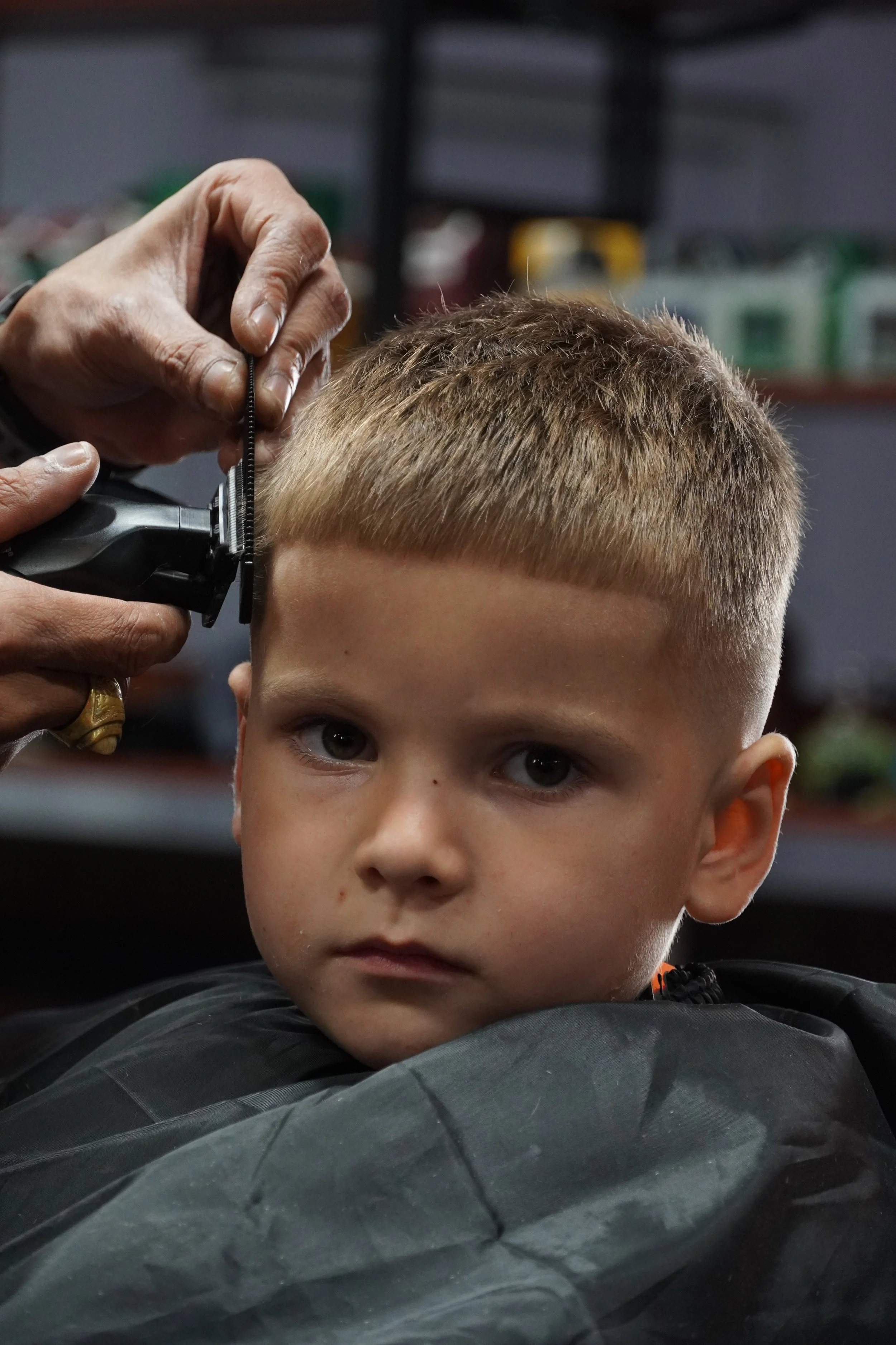 A young boy with short blond hair getting a haircut at a barber shop, with a barber trimming his hair.