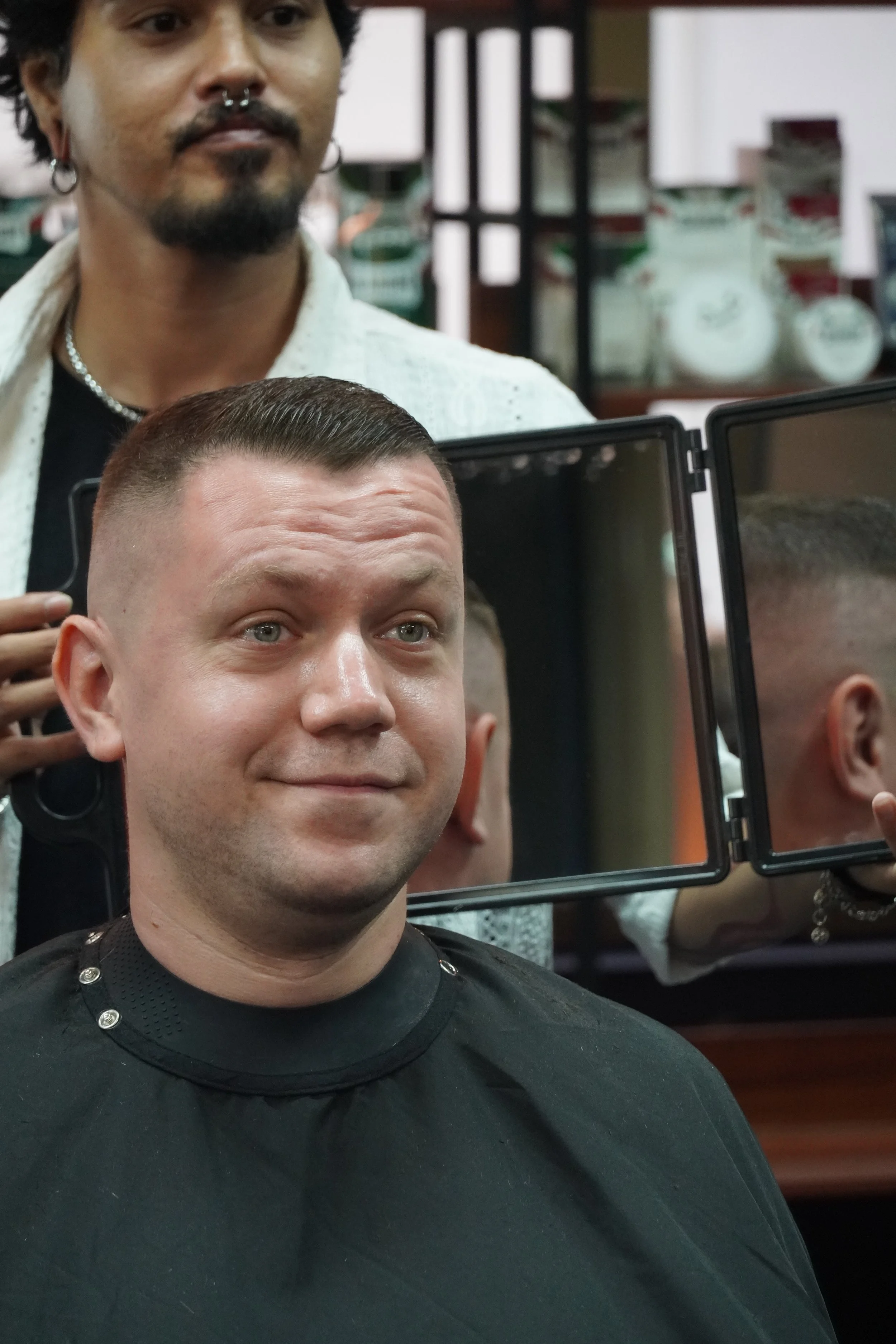 A man with a buzz cut sitting in a barber chair, looking at his reflection in the mirror. A barber with dark curly hair and piercings stands behind him in a barbershop.