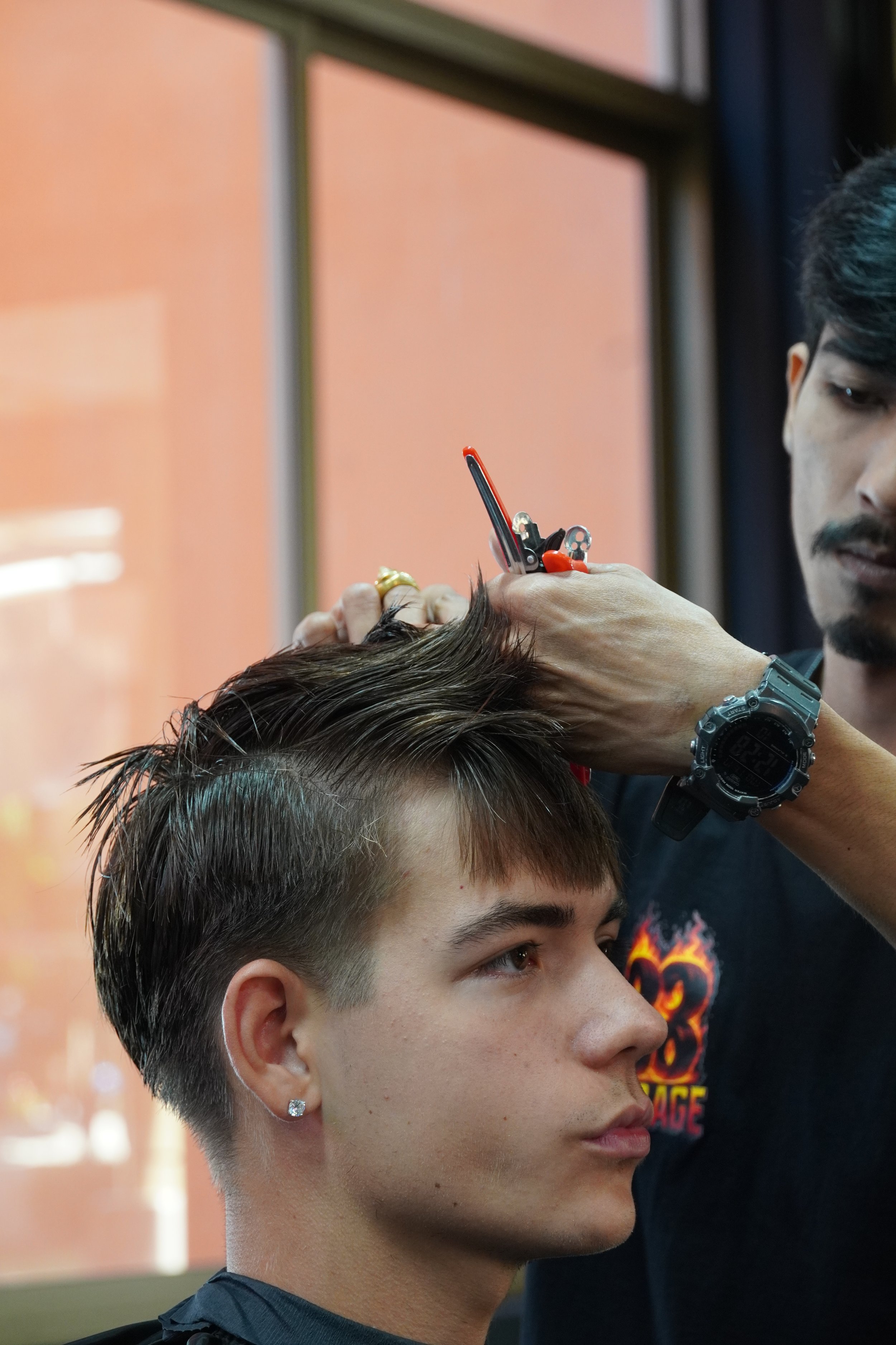 A barber is giving a haircut to a young man with dark hair, wearing a black shirt and earrings, in a barbershop.