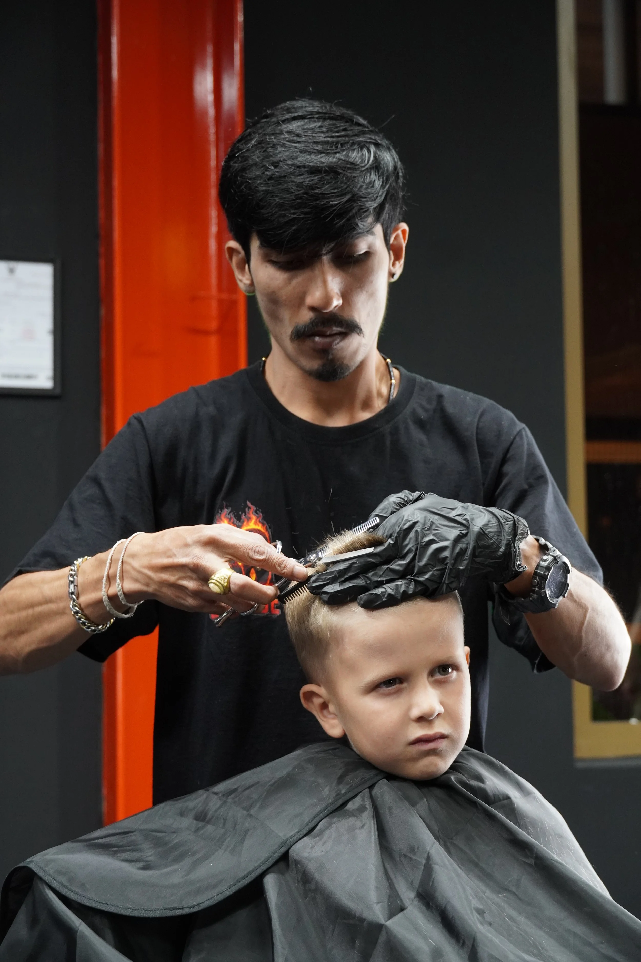 A young boy with blonde hair gets a haircut from a male barber with dark hair and tattoos. The barber is wearing black gloves and haircutting tools, and both are focused on the haircut.