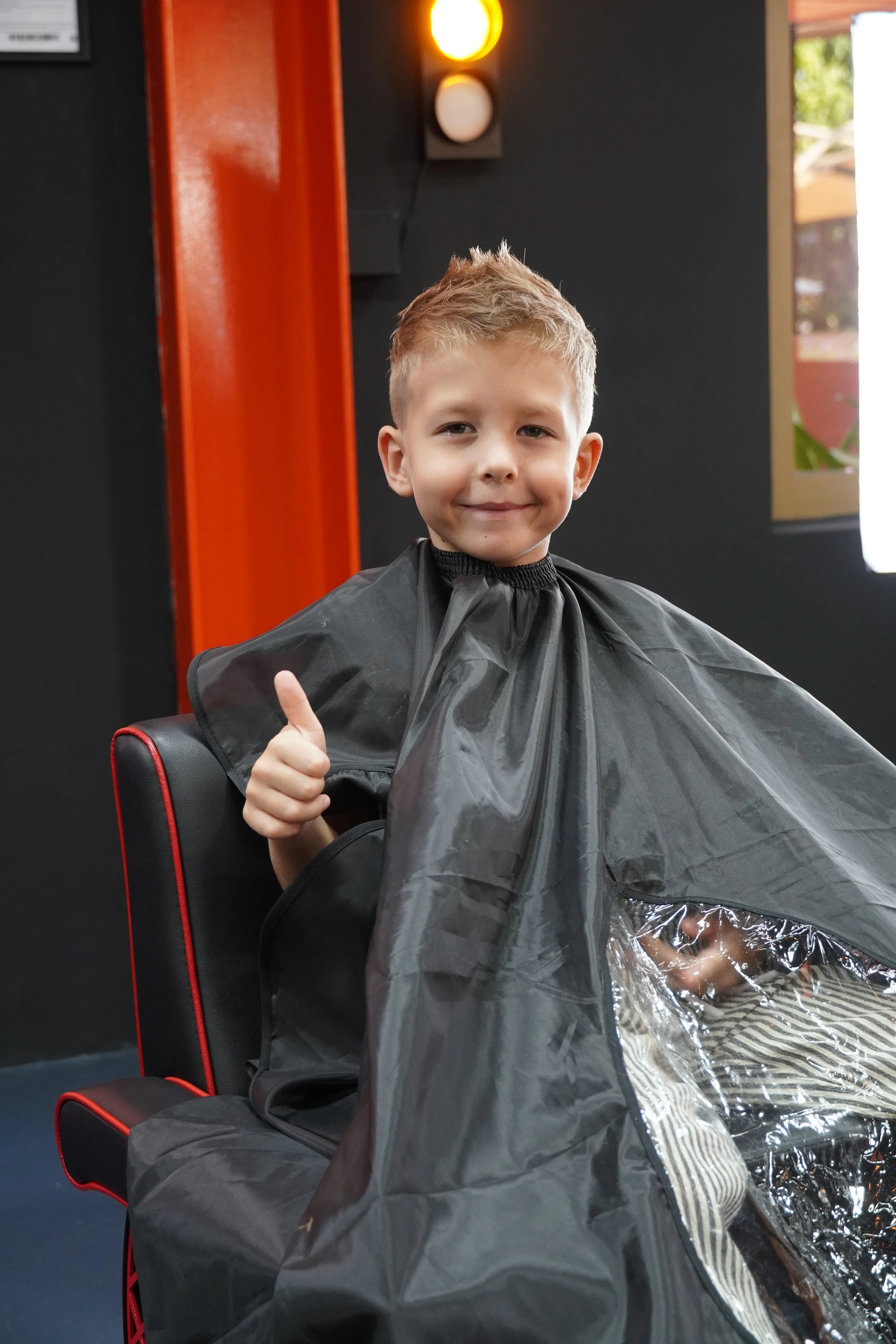 A young boy with light brown hair giving a thumbs up while sitting in a chair at a hair salon, wearing a black cape and having his hair cut.