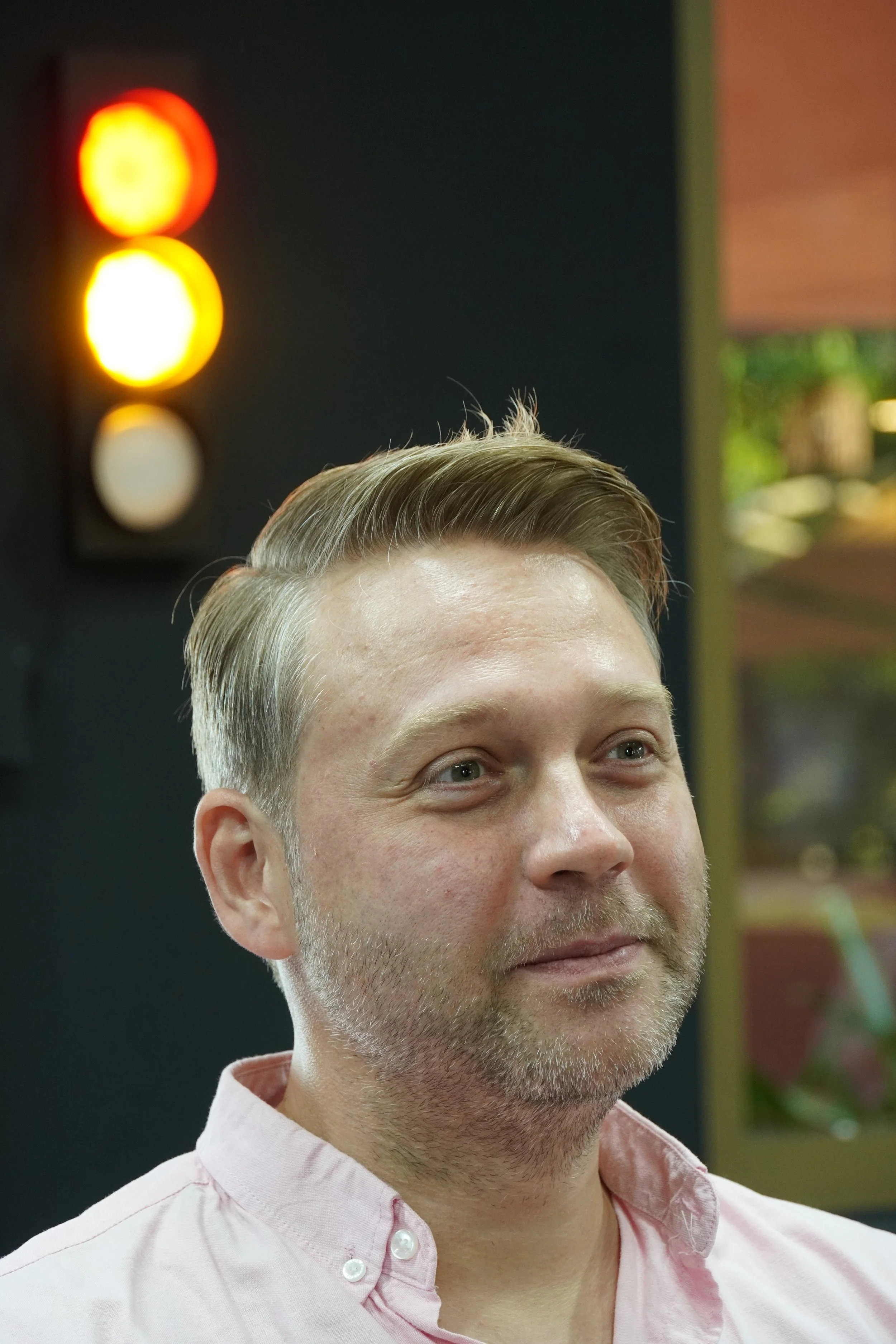 A man with a beard and short, light brown hair, wearing a light pink button-up shirt, standing indoors near a traffic light showing red, yellow, and white signals.