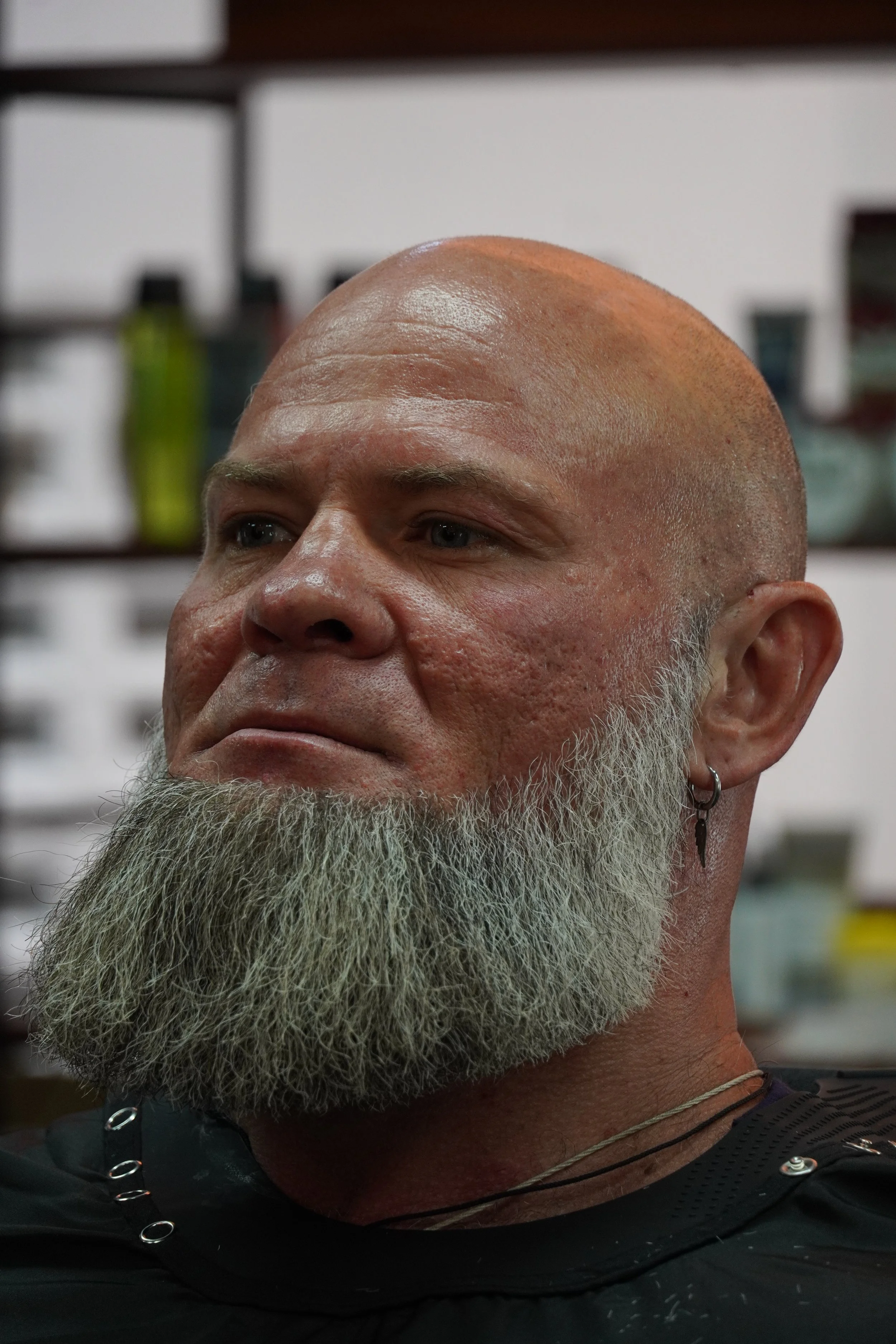 Close-up of a bald man with a gray beard, wearing a black shirt, earrings, and a necklace, in an indoor setting.
