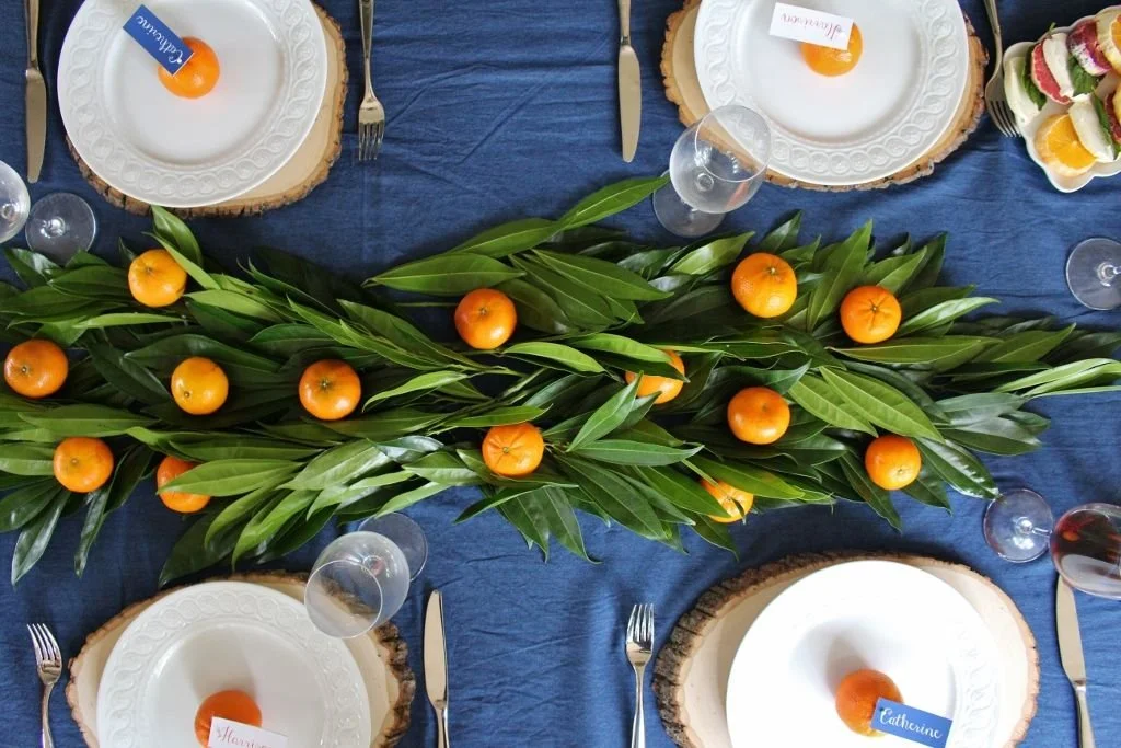 A table decorated with green leaves and small orange fruits, with white plates, silverware, and glasses set for a meal.