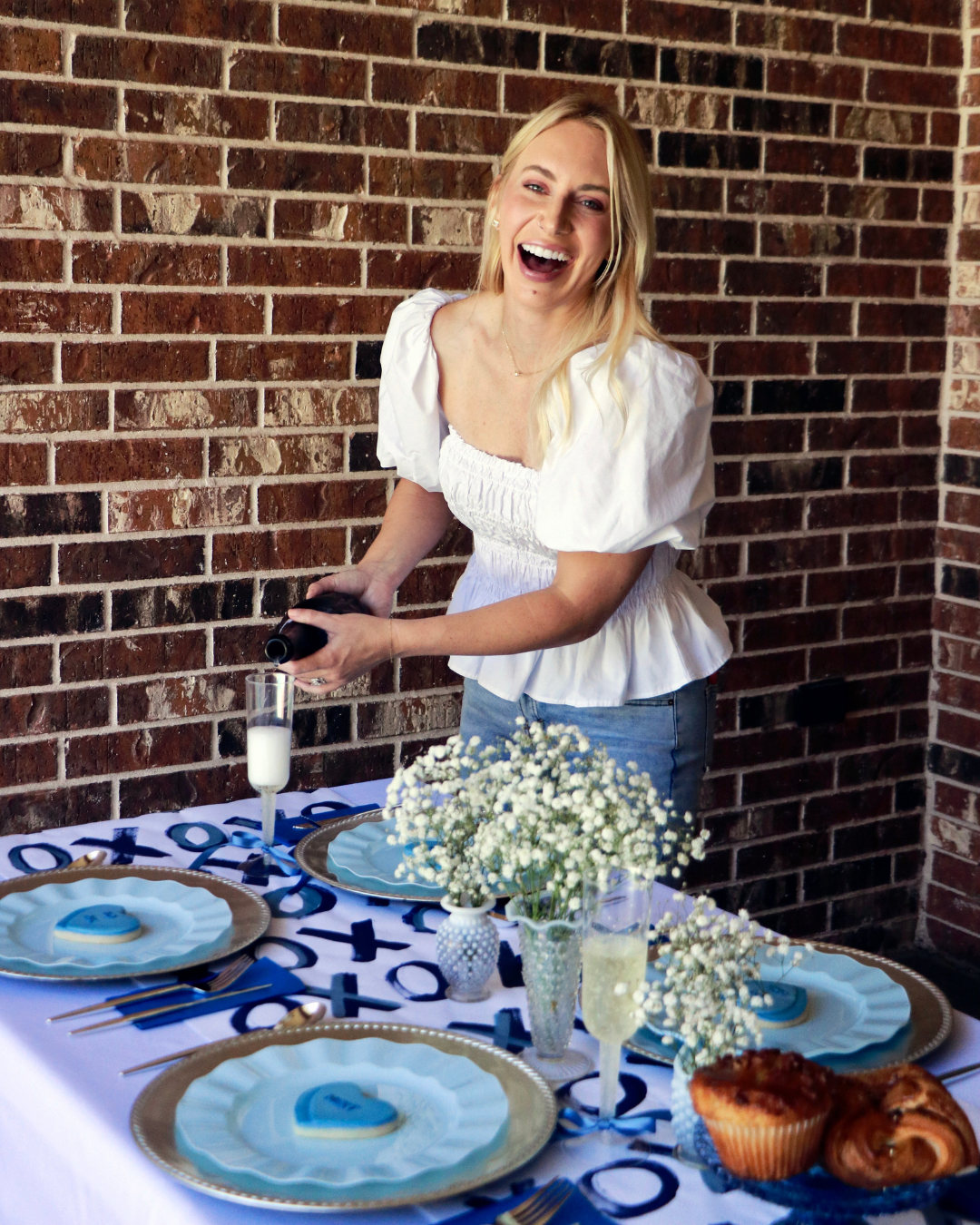 A woman smiling and pouring champagne at a decorated table with blue and white tableware, flowers, and pastries, in front of a brick wall.