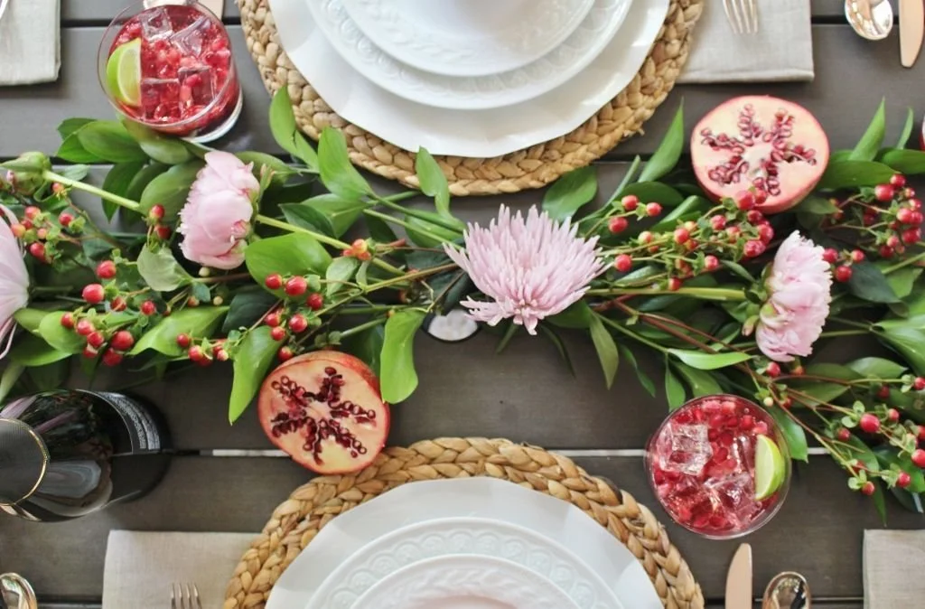 A table setting with floral decorations and refreshments. There are floral and berry garlands with pink flowers and red berries running down the center of the table, along with slices of pomegranate and a pinkish cake decorated with berries. Place settings with white plates on woven placemats are arranged around the table.