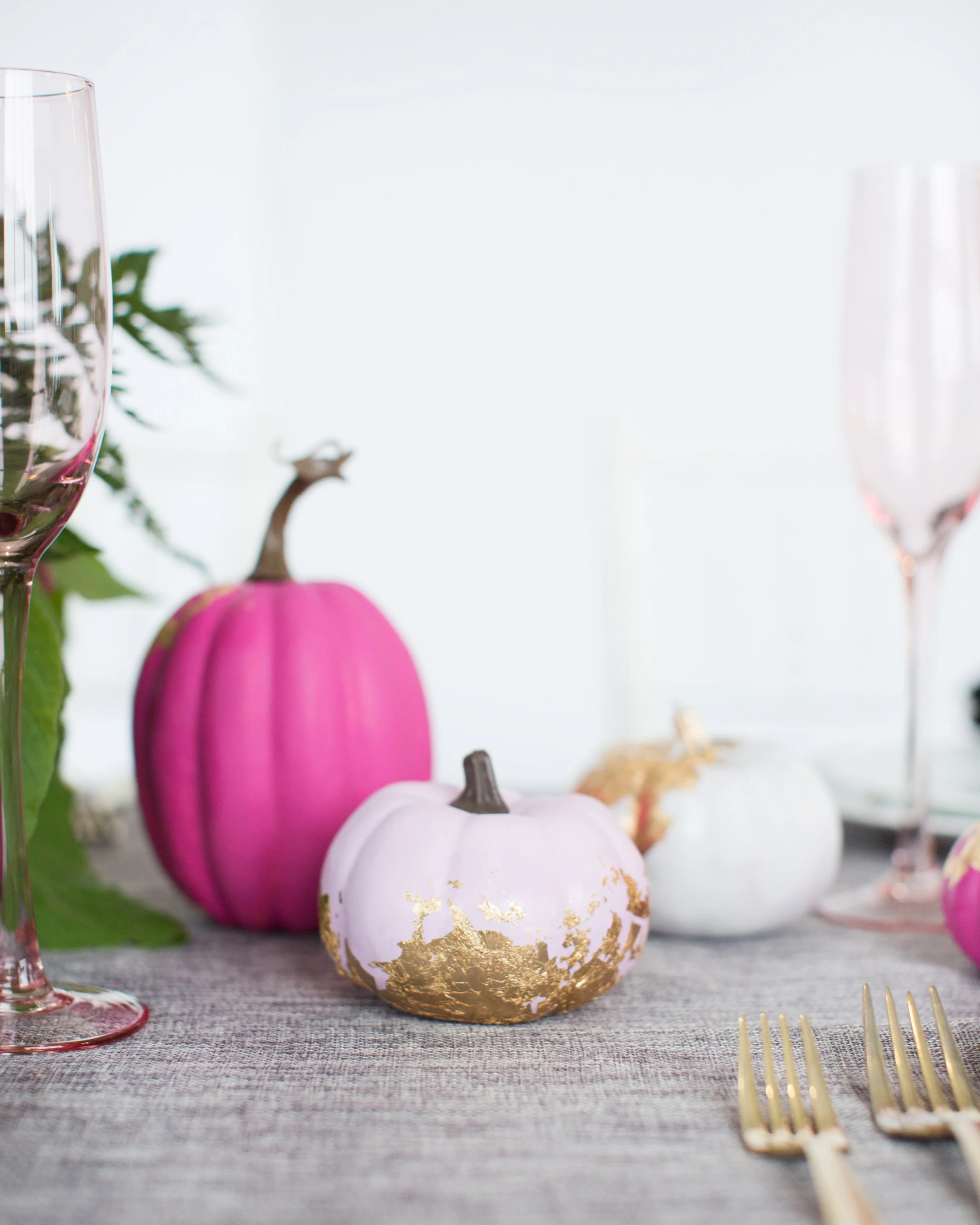 Decorative pumpkins on a table, with pink, white, and gold highlights, alongside glassware and gold utensils, with greenery in the background.