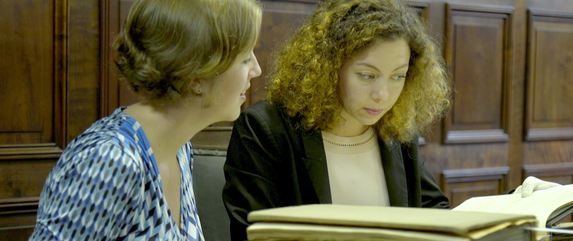 Screenshot from a movie with two women sitting at a table in a room with wooden paneling, looking at a large open book.