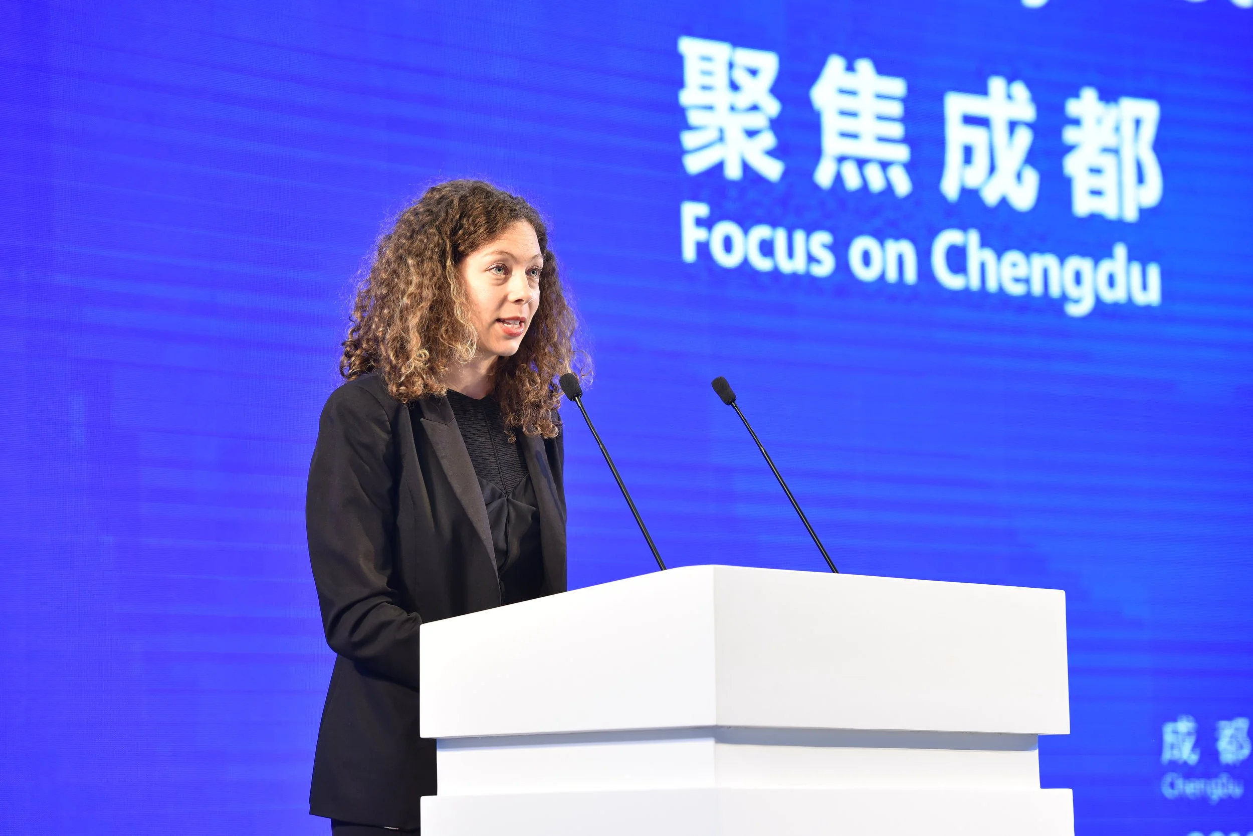 A woman with curly hair wearing a black blazer and black top, speaking at a white podium with two microphones, in front of a large blue screen with Chinese characters and the English words "Focus on Chengdu."
