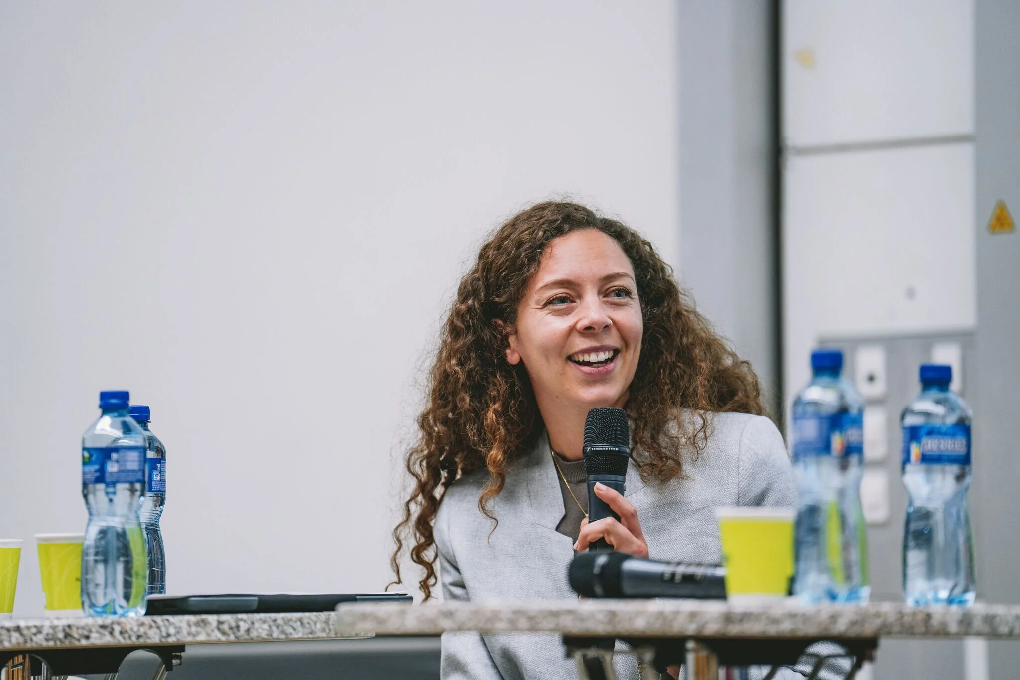 A woman with curly hair holding a microphone, smiling, sitting at a table with water bottles and yellow cups, in a conference or meeting room.