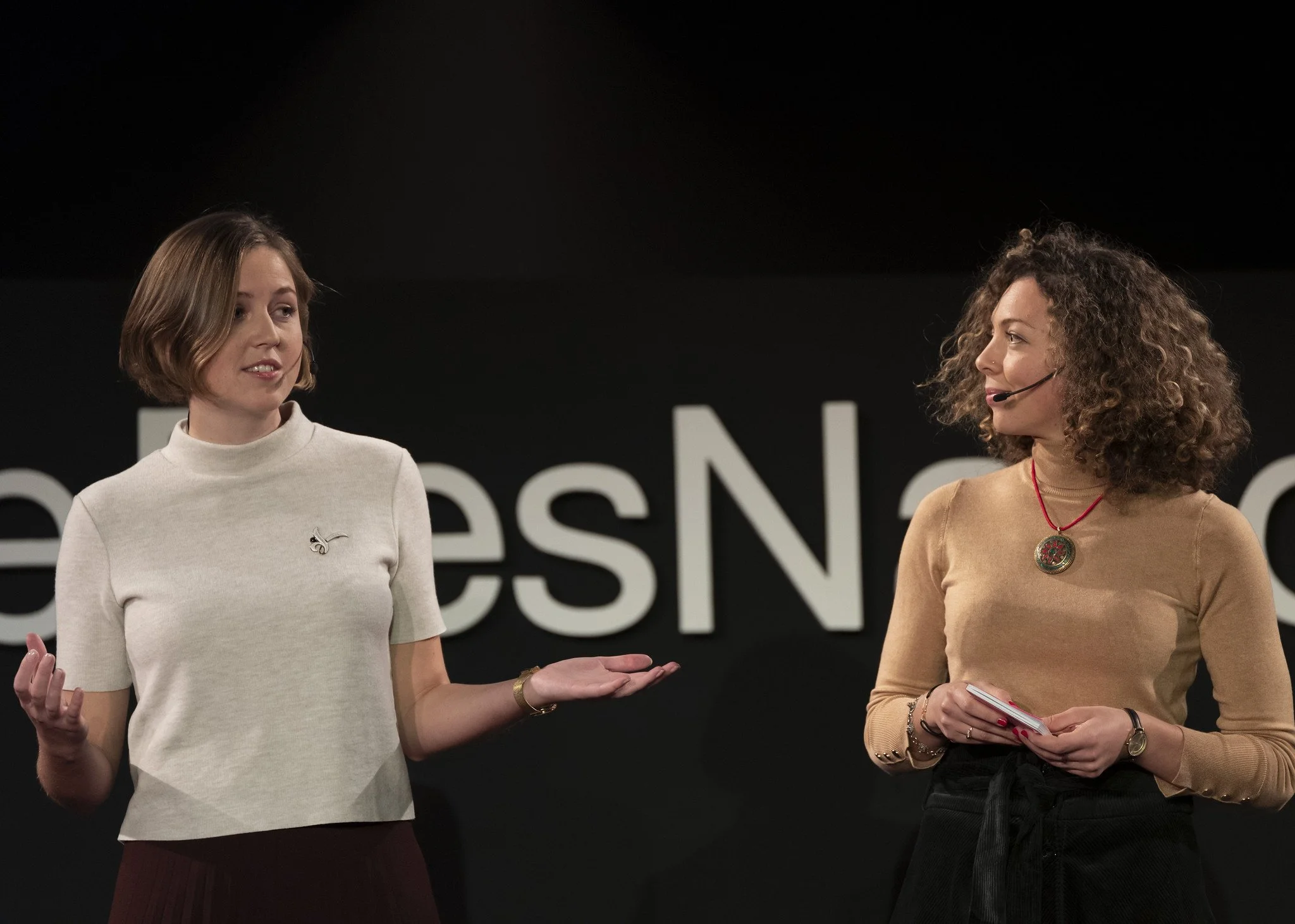 Two women are on stage, engaged in a conversation during a presentation or talk. The woman on the left has short hair, wears a cream-colored top with a small emblem and a burgundy skirt. The woman on the right has curly hair, wears a tan top with a r