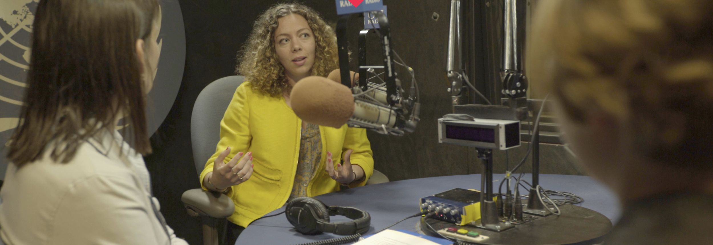A woman in a yellow jacket is speaking into a microphone during a radio interview at a recording studio.