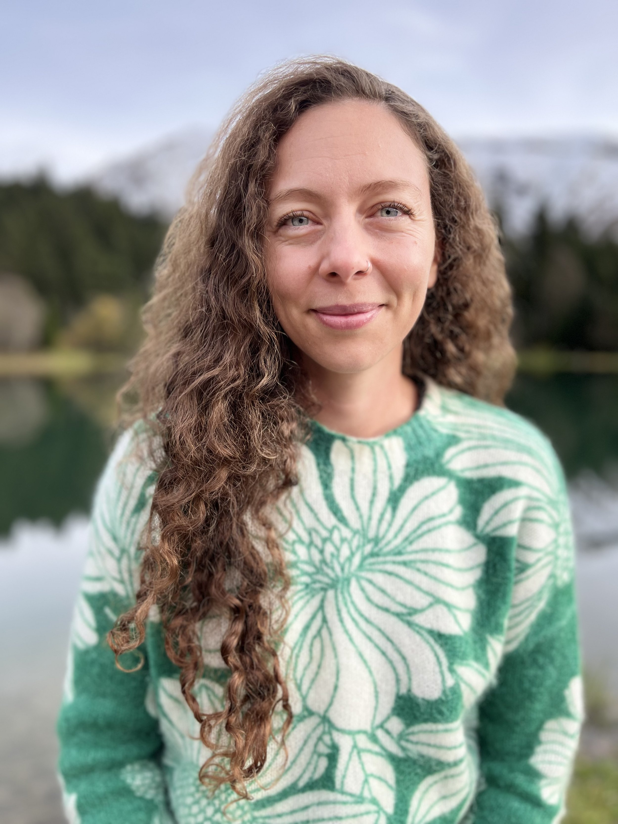A woman with curly brown hair and blue eyes smiling outdoors with a blurred mountain and water background, wearing a green and white floral sweater.