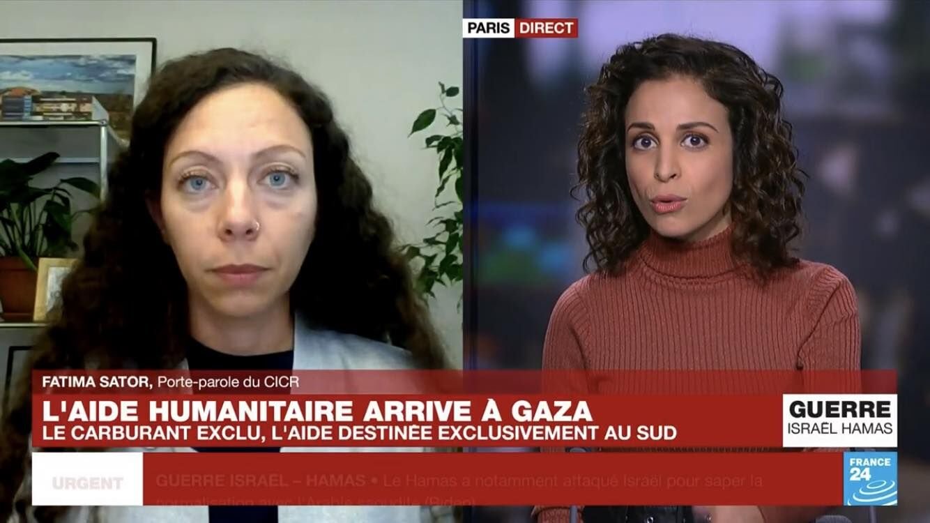 Split-screen image showing two women during a news interview. The woman on the left has long, curly brown hair, light skin, and is in a room with plants and framed artwork. The woman on the right has dark curly hair, medium skin, and is wearing a rust-colored turtleneck sweater, with a blurred background.