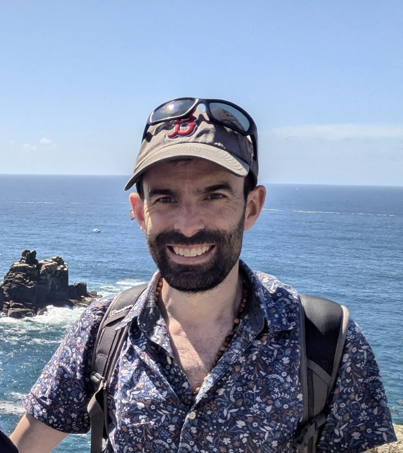 A smiling man with a beard and dark hair wearing a beige cap, sunglasses, a patterned shirt, and a black backpack stands outdoors near the ocean with rocks and blue sky in the background.