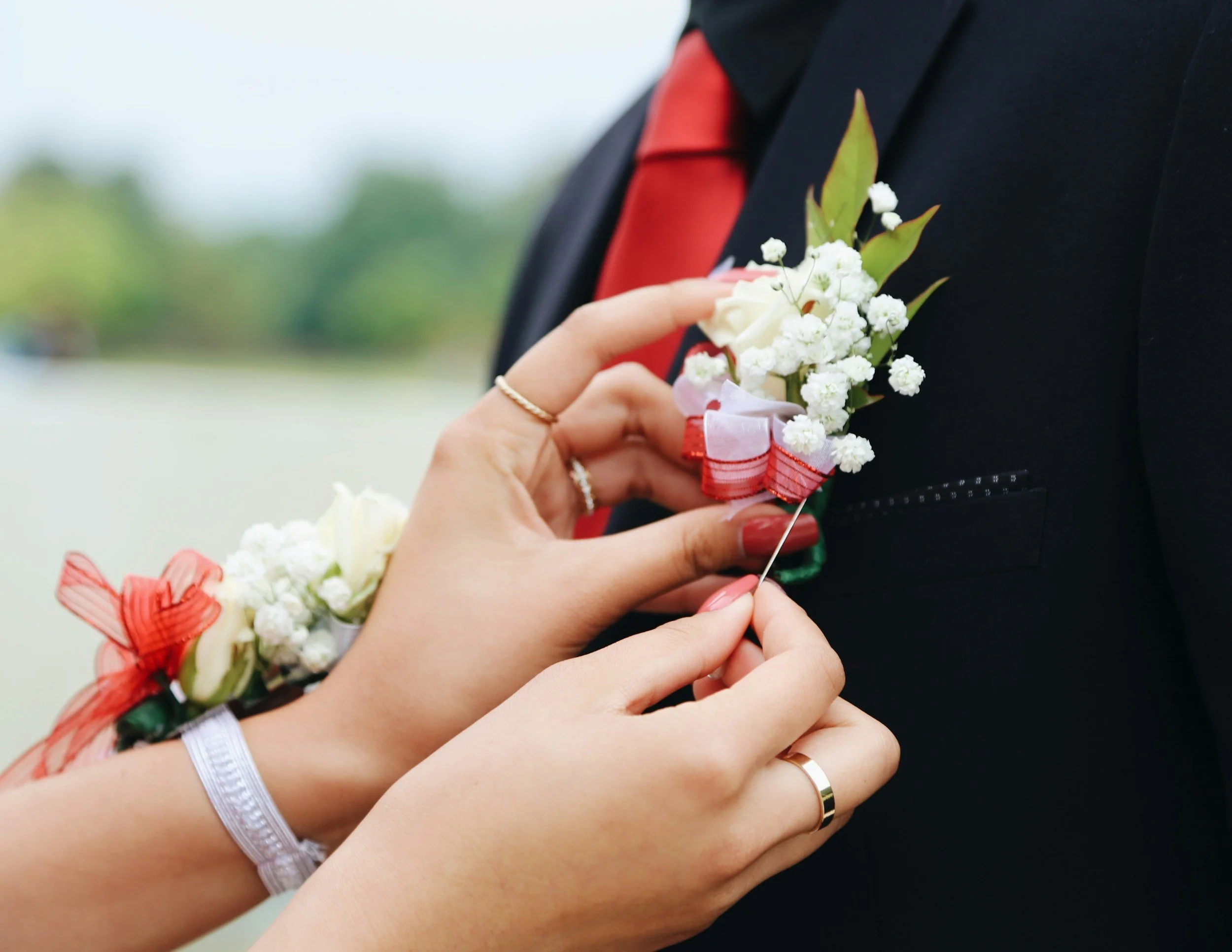 Close-up of a person pinning a boutonniere with white flowers, green leaves, and a red ribbon on a man's black suit jacket during a wedding.