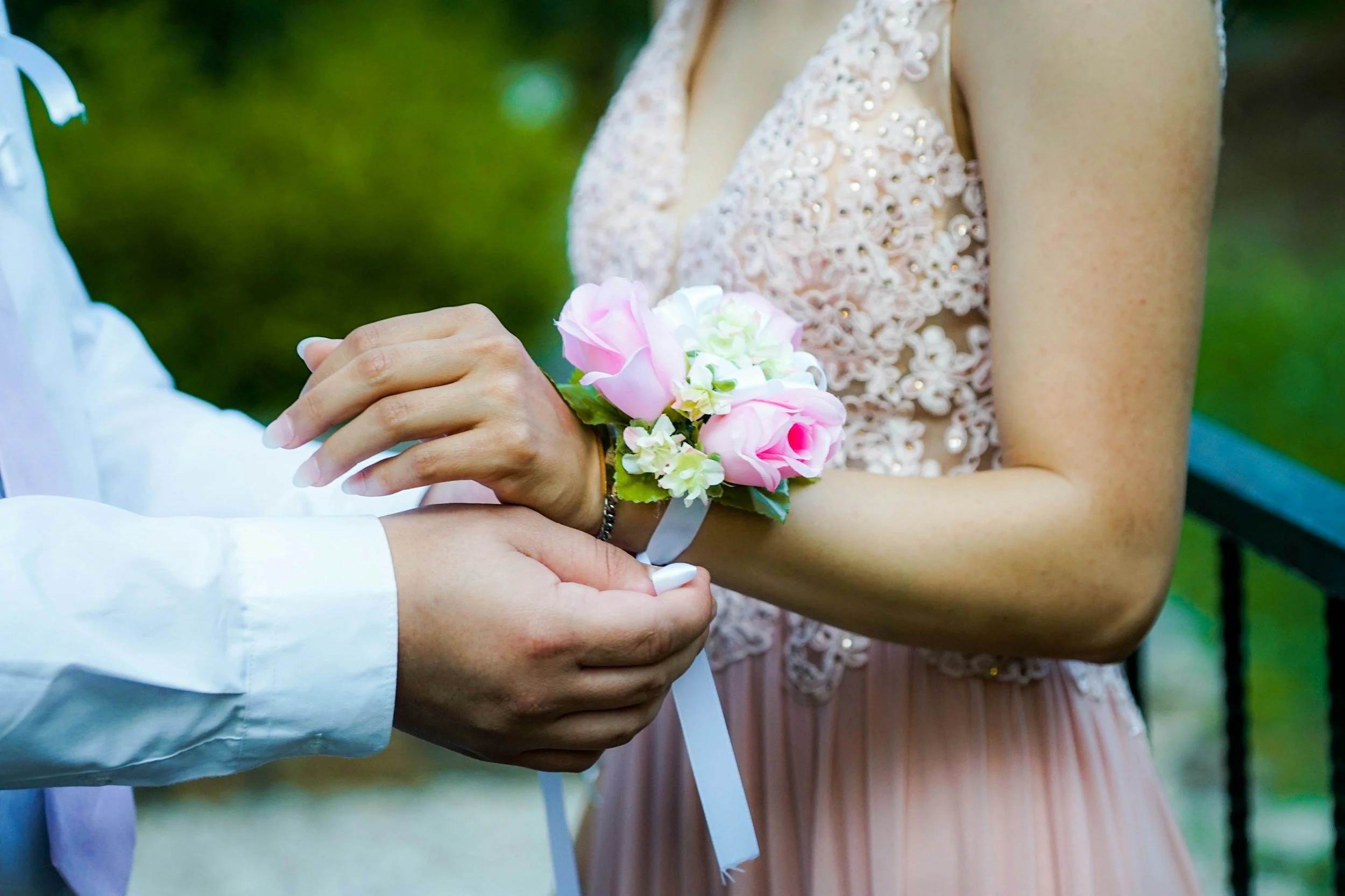 A close-up of a couple holding hands during a wedding ceremony, with the bride wearing a floral wrist corsage and a detailed lace dress, and the groom dressed in a white shirt.