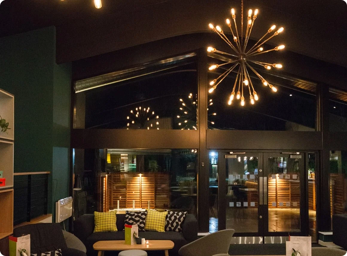 Interior of a modern lounge or restaurant with a dark ceiling, large glass doors, a chandelier with multiple exposed bulbs, and a seating area decorated with black and yellow cushions.
