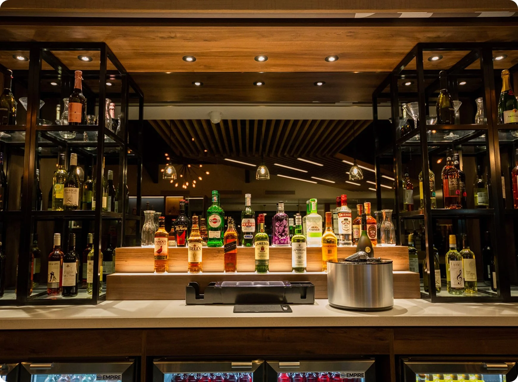 Bar with a variety of liquor bottles displayed on shelves, illuminated by warm ceiling lights, with a modern design featuring wooden and metal accents.