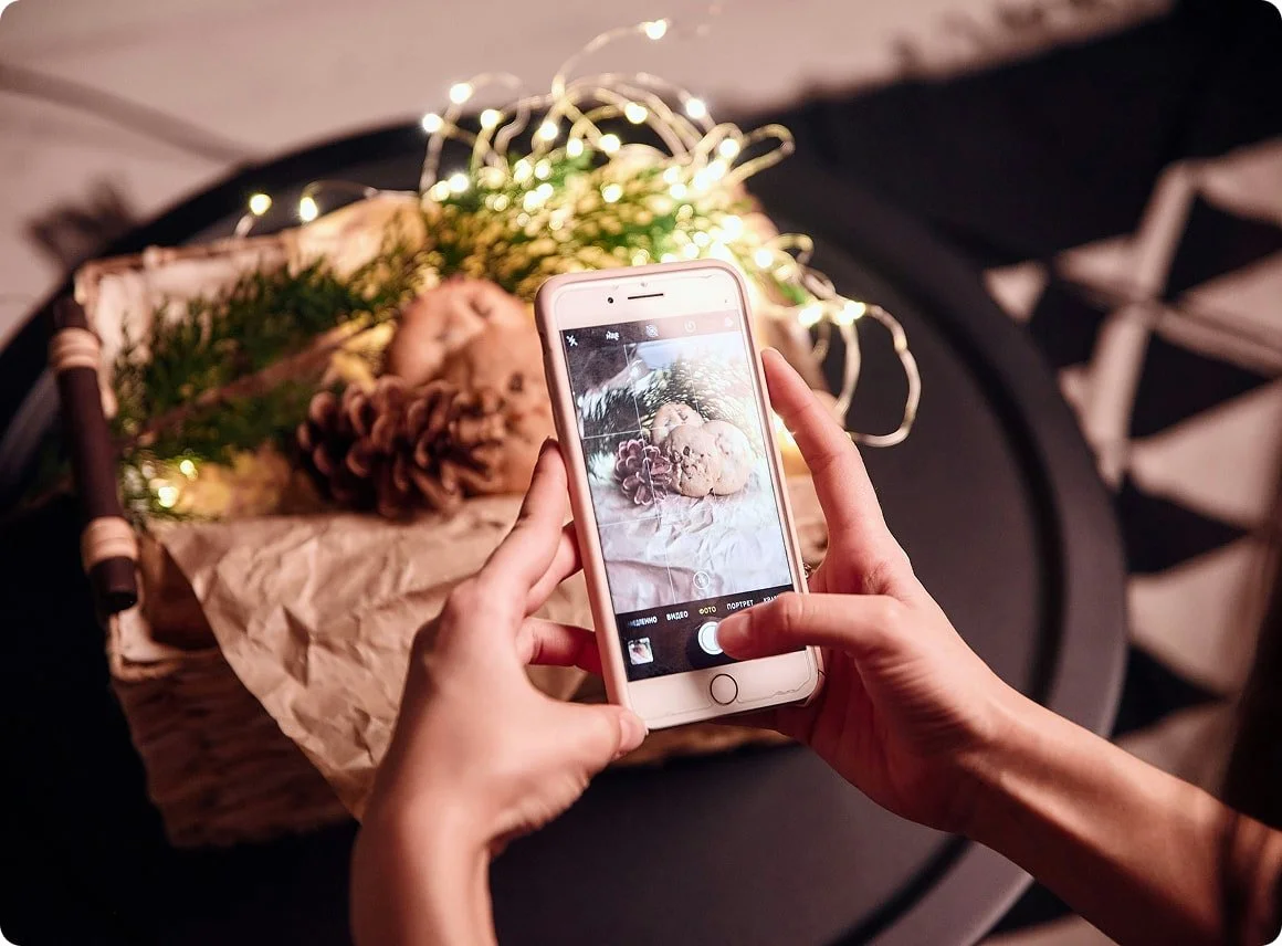 Person taking a photo of a gift box decorated with pine cones, greenery, and string lights with a smartphone.