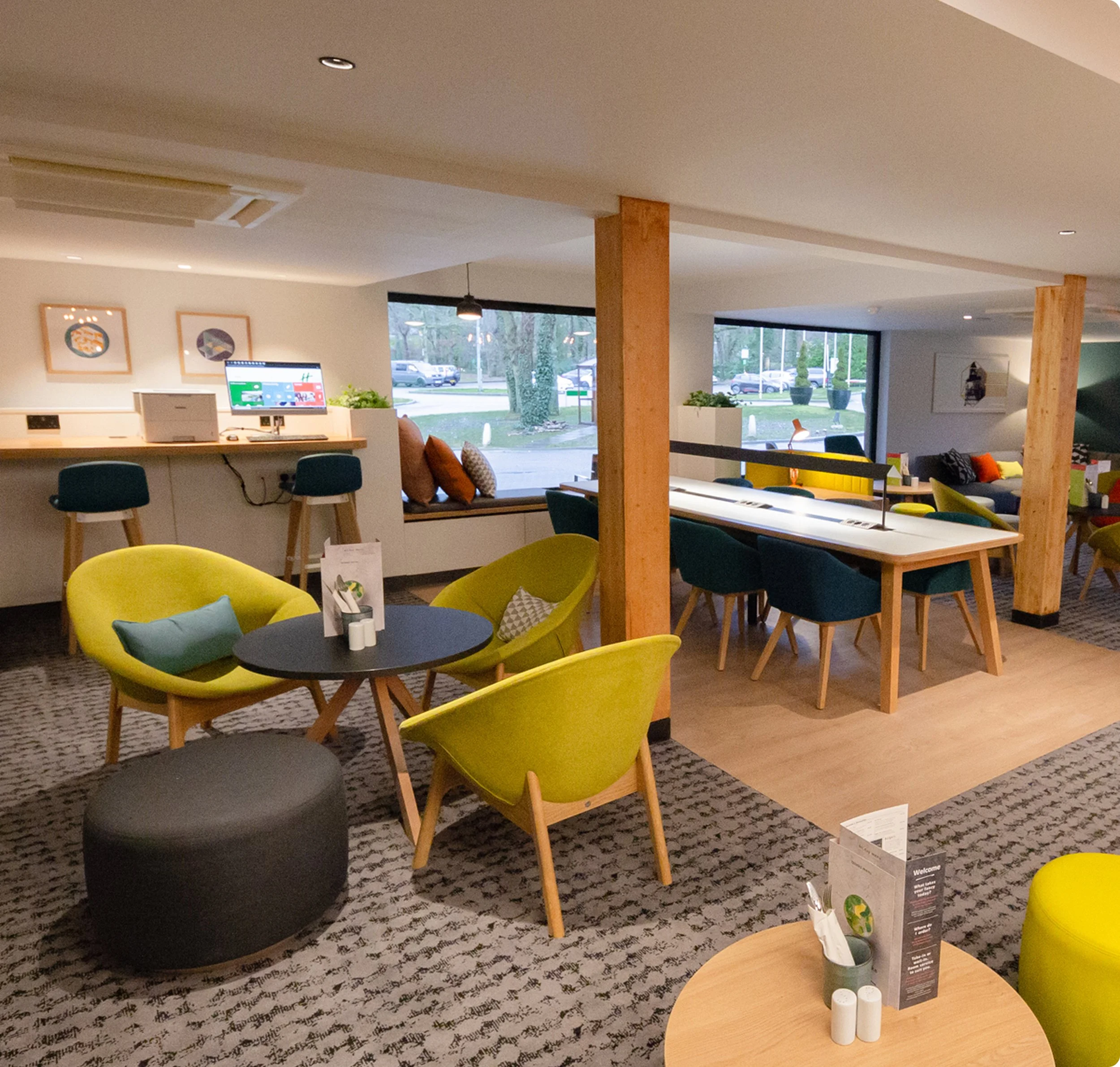 Interior view of a modern lounge or waiting area with colorful chairs, a counter with a monitor, framed artwork on the wall, and large windows showing parked cars outdoors.