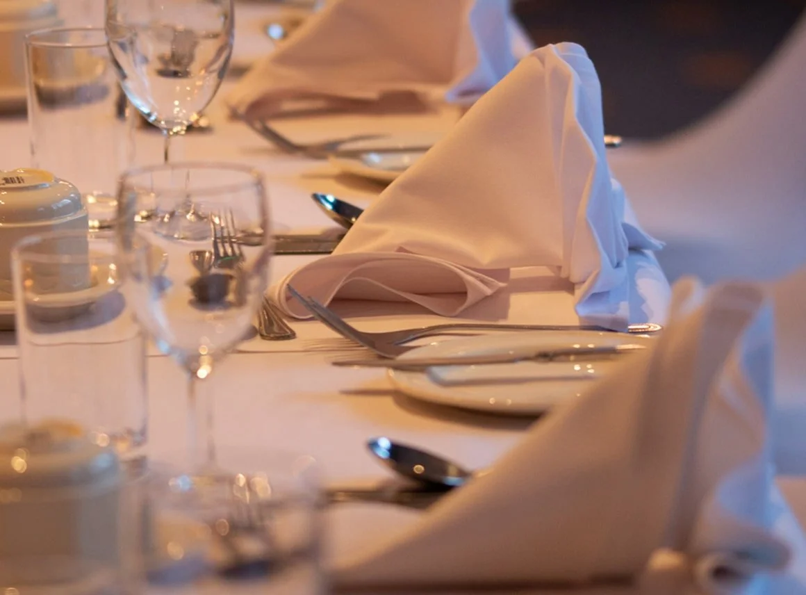 A neatly set dining table with white napkins folded into a tall, pointed shape, multiple glasses, silverware, and a white tablecloth, prepared for a formal event or dinner.