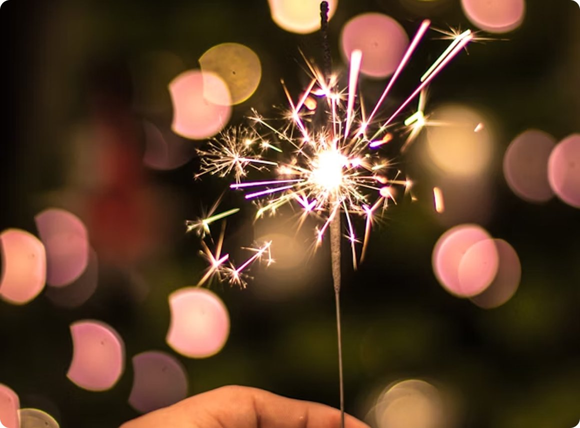 A lit sparkler creating bright sparks against a blurred background with pink and yellow bokeh lights.