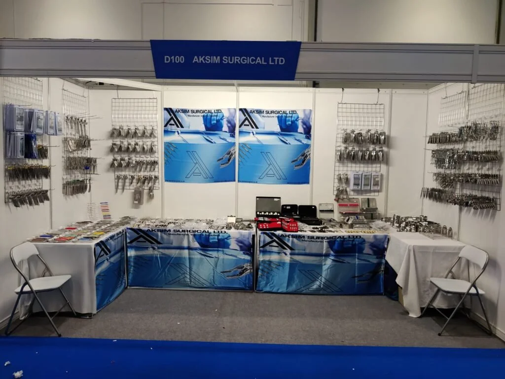 Exhibit booth for Aksim Surgical Ltd displaying various surgical instruments and supplies, with two foldable chairs and promotional banners.