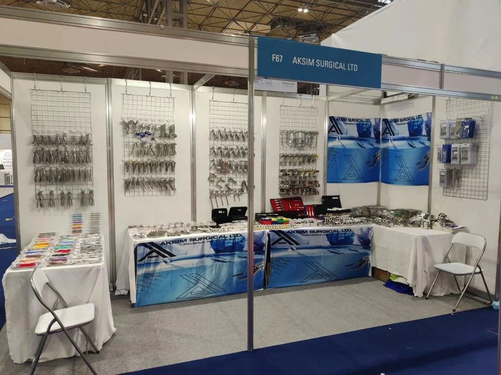 Trade show booth displaying various surgical instruments and medical supplies from AKSIM SURGICAL LTD, with walls of hooks holding instruments and tables of supplies.