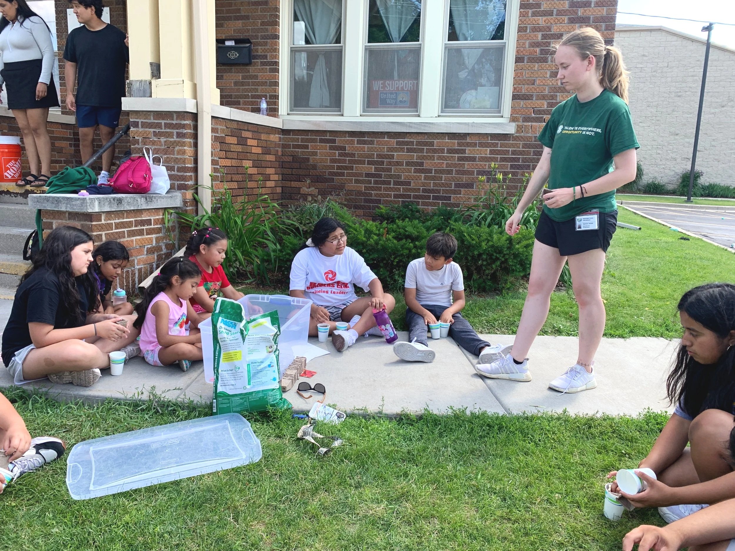 A group of young children sitting on the sidewalk and grass, participating in an outdoor activity, with an adult standing nearby. The scene appears to be at a school or community center, with some supplies and snacks in view.
