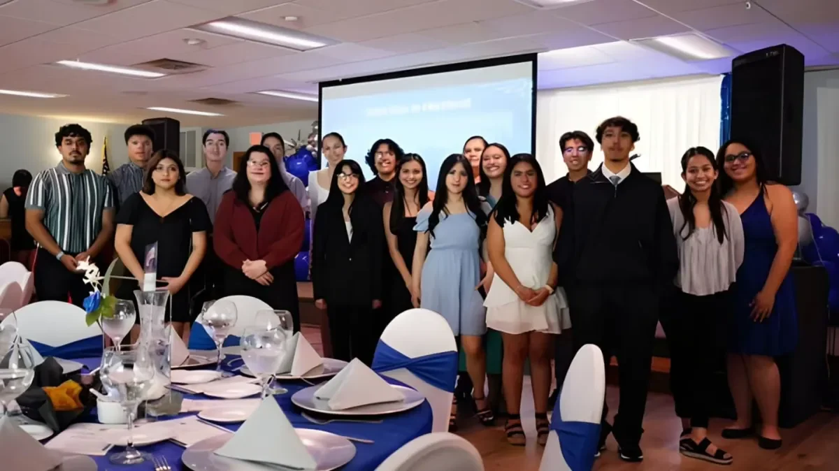 Group of young adults in formal attire posing for a photo at a celebration event with decorated tables and a projector screen in the background.