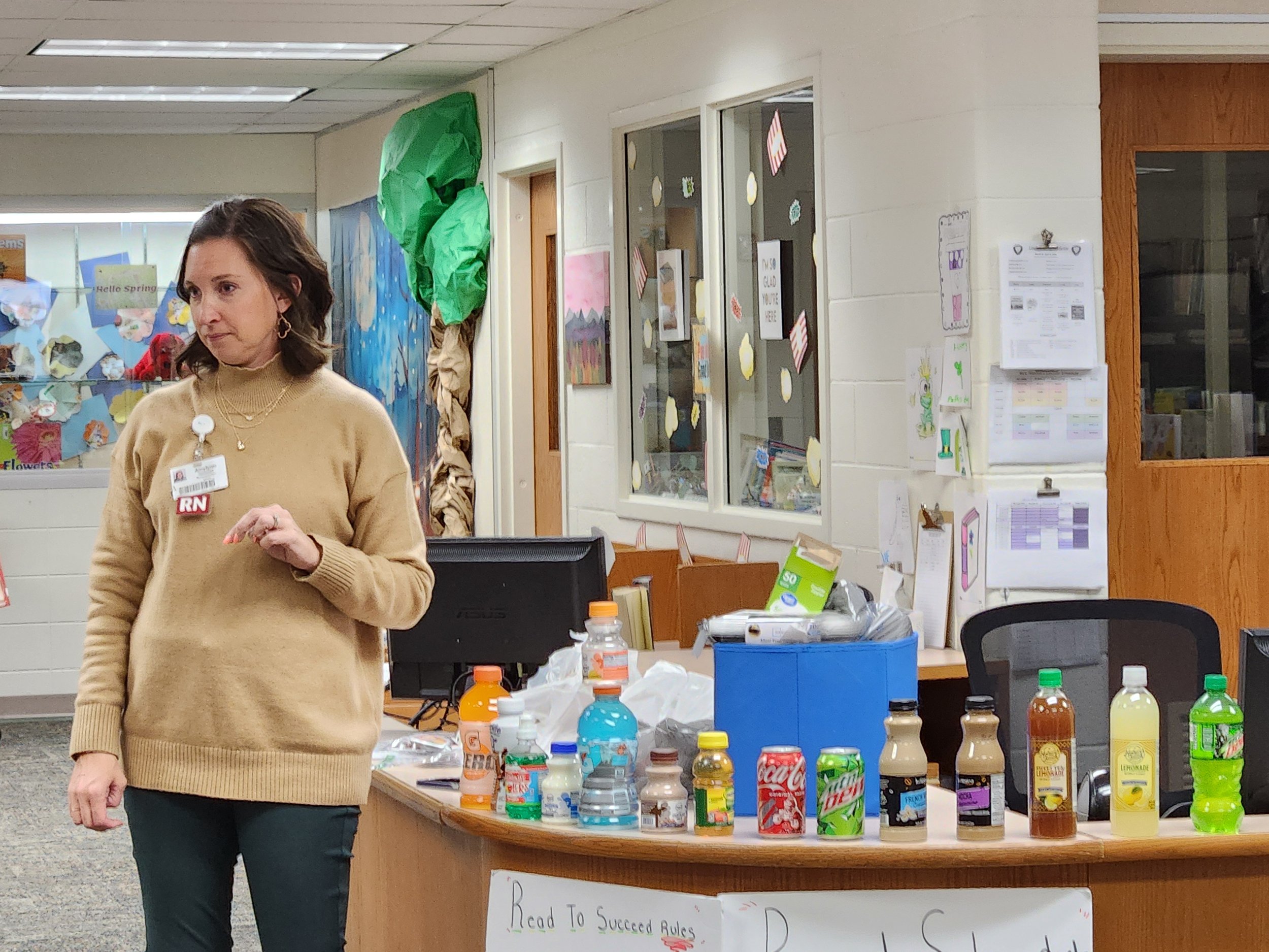 A woman wearing a beige sweater with an RN badge stands next to a desk with beverages and a computer in a classroom or office setting, with colorful drawings and papers posted on the walls.
