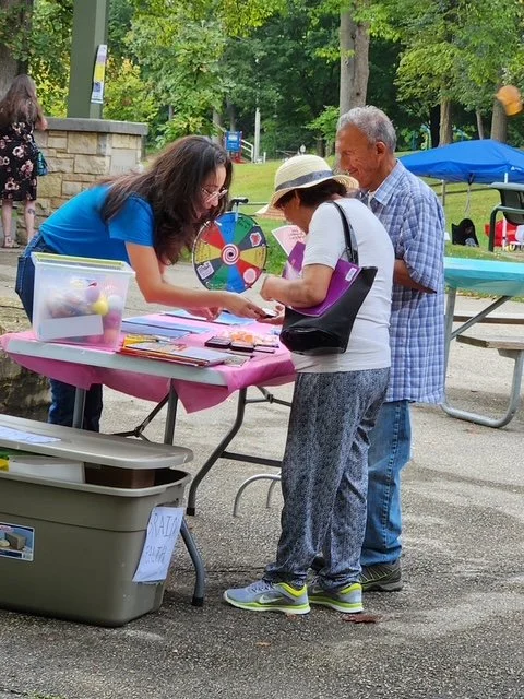 Three people at an outdoor event near a pink table with colorful items, a spinning wheel, and a clear plastic box, with trees and other booths in the background.