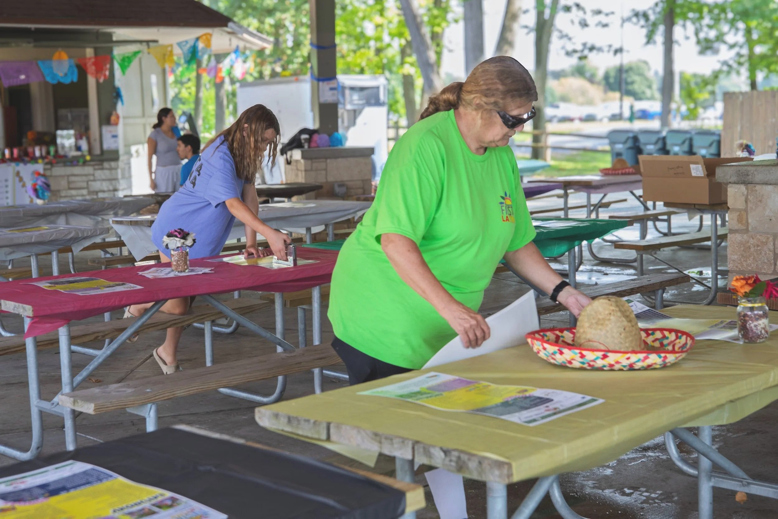 People setting up booths with tables and decorations at an outdoor event under a pavilion with trees in the background.