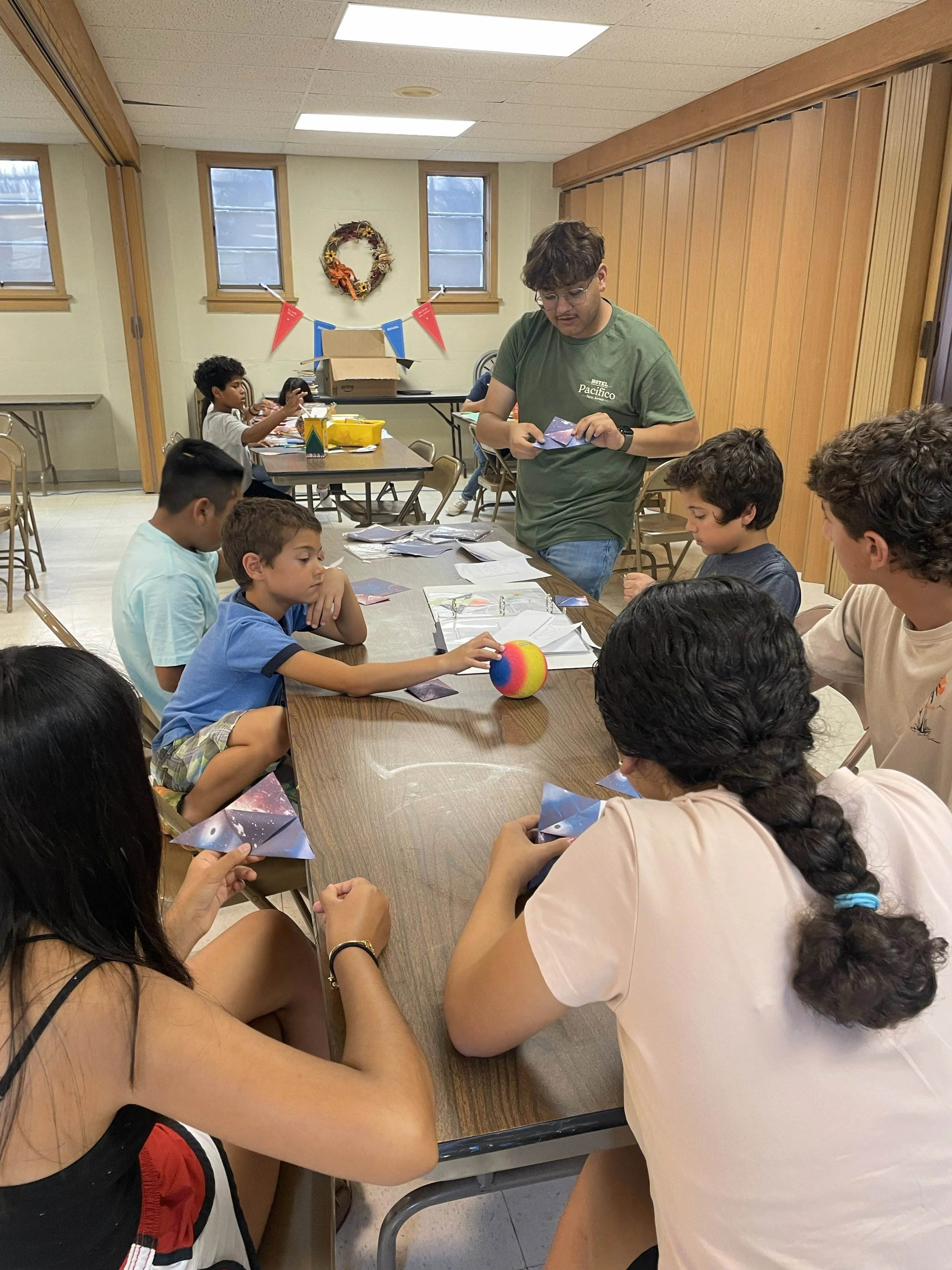 Children and adults sitting around a table engaging in an activity involving space-themed cards and a rainbow-colored ball, with a person standing and observing.