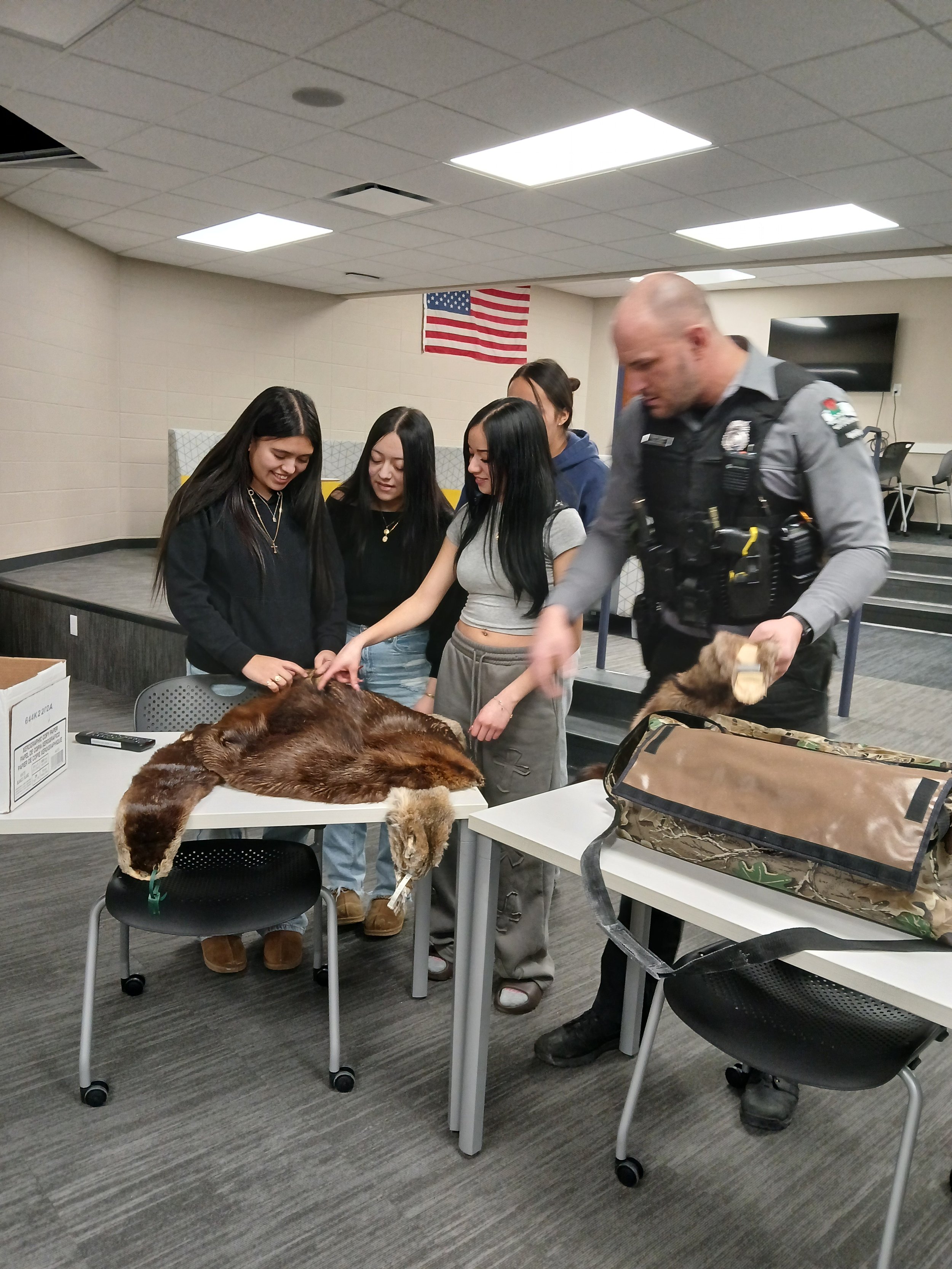 Group of young women and a police officer gathered around a table with a large, stretched-out fur of an animal, possibly a bobcat or lynx.