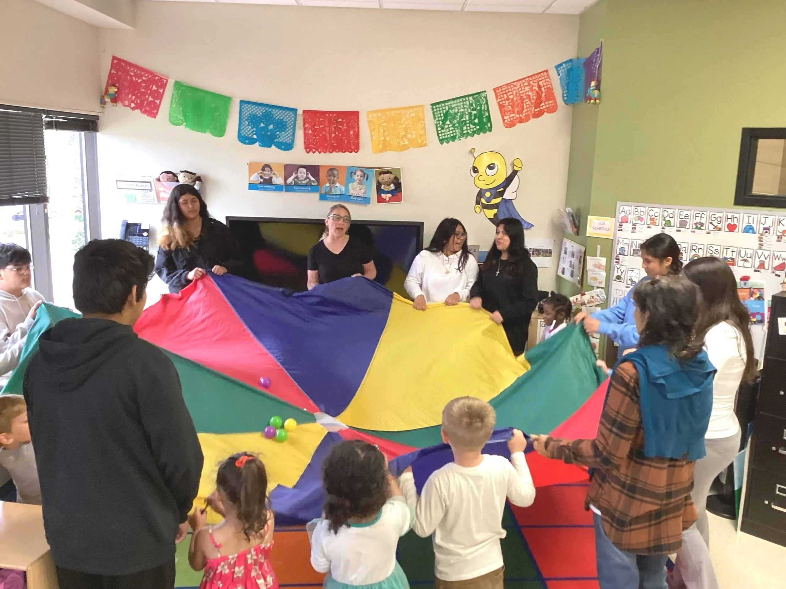Group of children and adults playing with a colorful parachute in a classroom decorated with banners and educational posters.