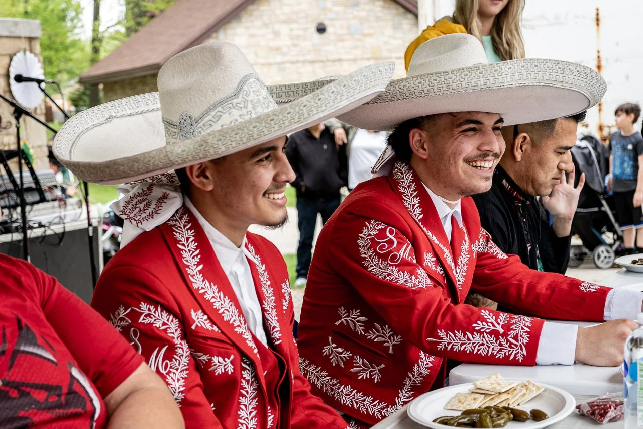 Three men wearing traditional Mexican mariachi suits and large sombreros sitting at a table outdoors, smiling.