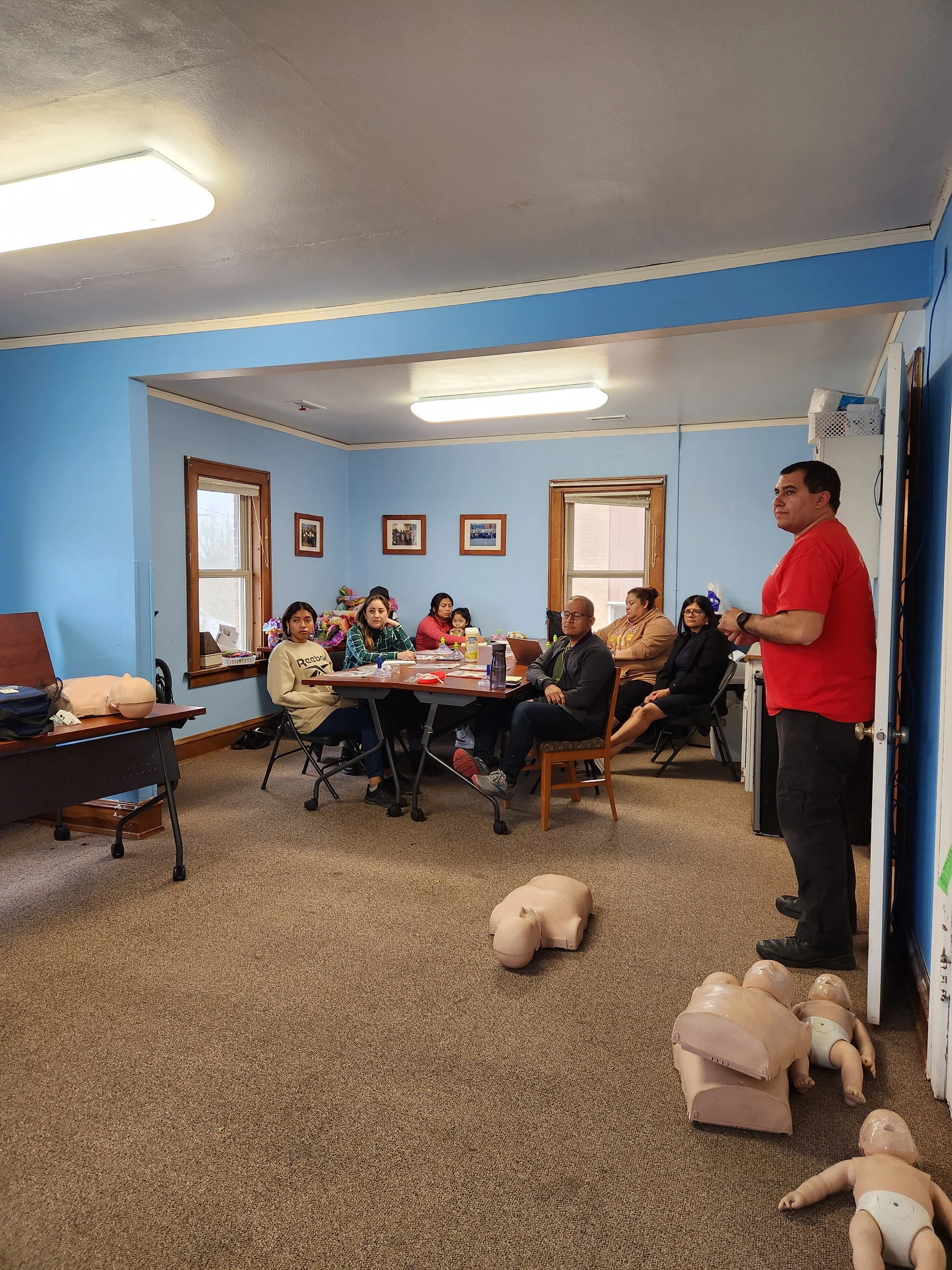 A man in a red shirt is standing in front of a group of people seated at a table in a classroom or training room. There are CPR mannequins on the floor near him. The room has blue walls, wooden window frames, and framed pictures on the wall.