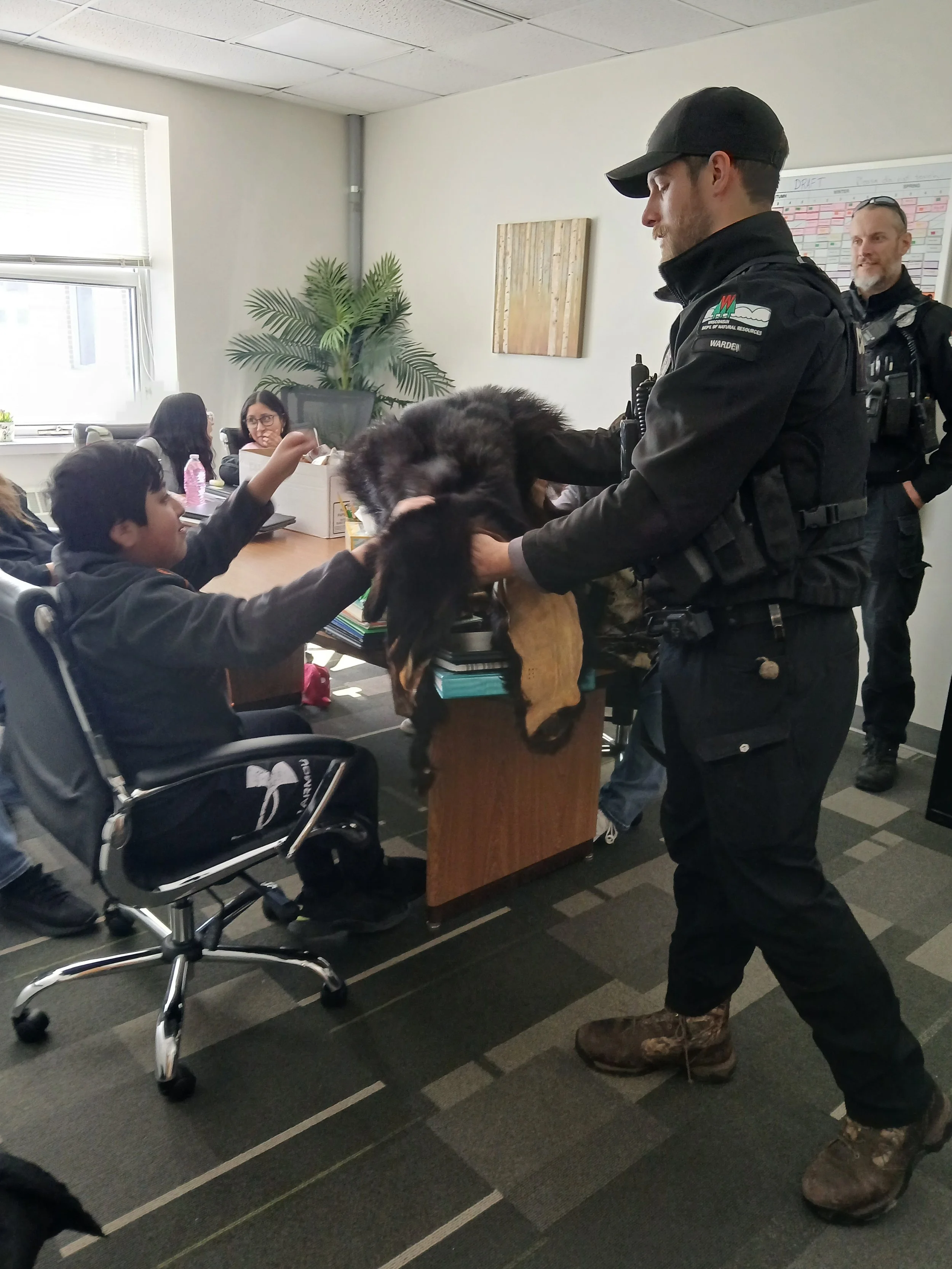 Police officer holding a large dog at a desk while children and adults watch and reach out to pet the dog in an office setting.