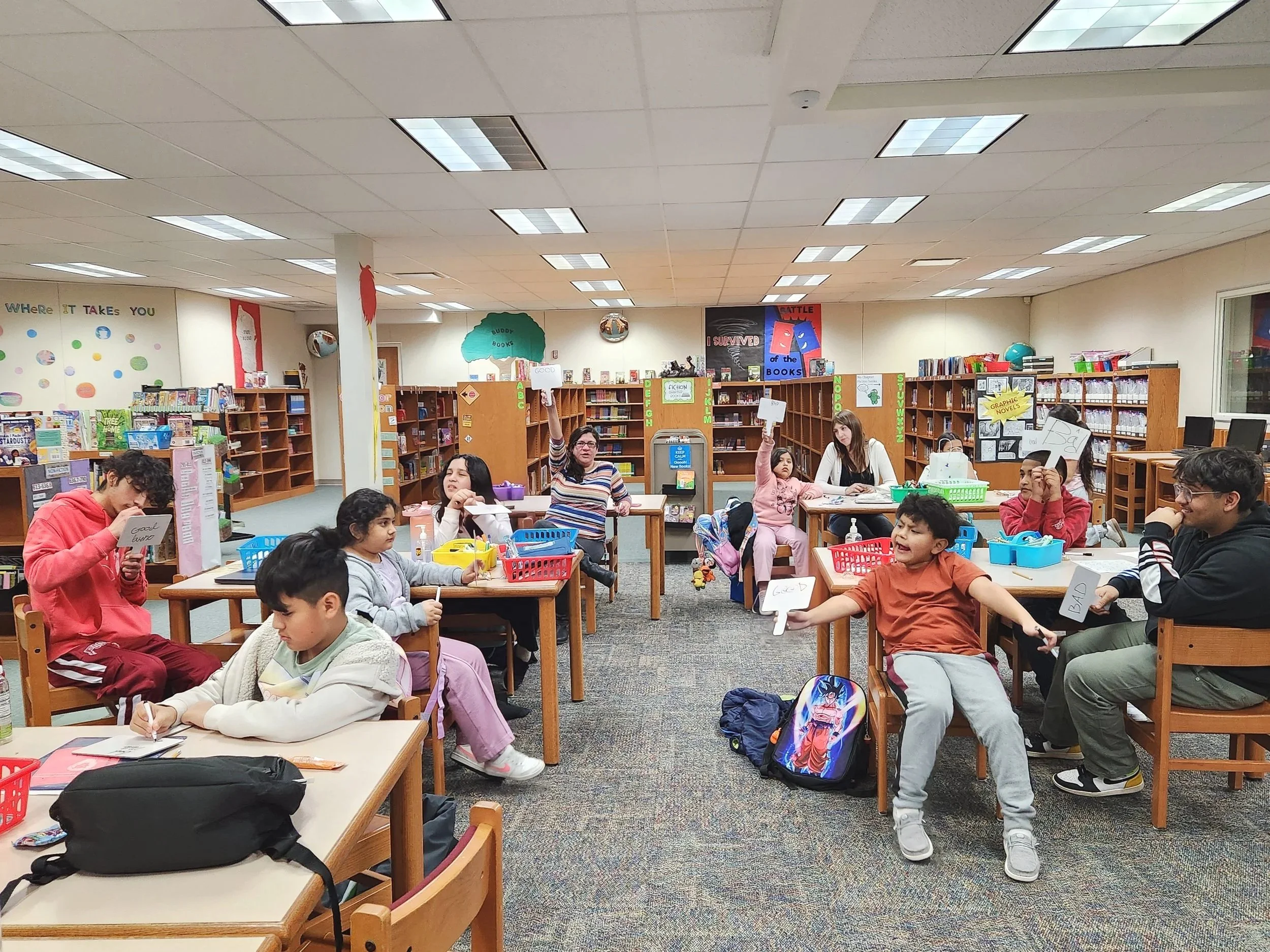 Students sitting at library tables, some raising whiteboards, in a school library surrounded by bookshelves and colorful decorations.