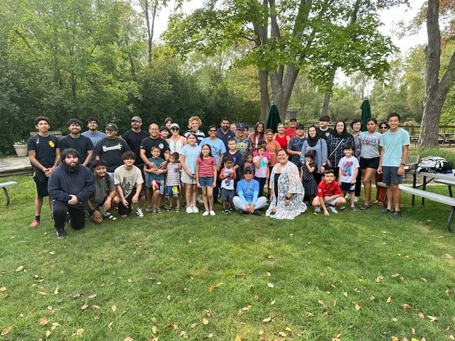 Group of people, including adults and children, gathered outdoors on a grassy area with trees in the background, posing for a group photo.