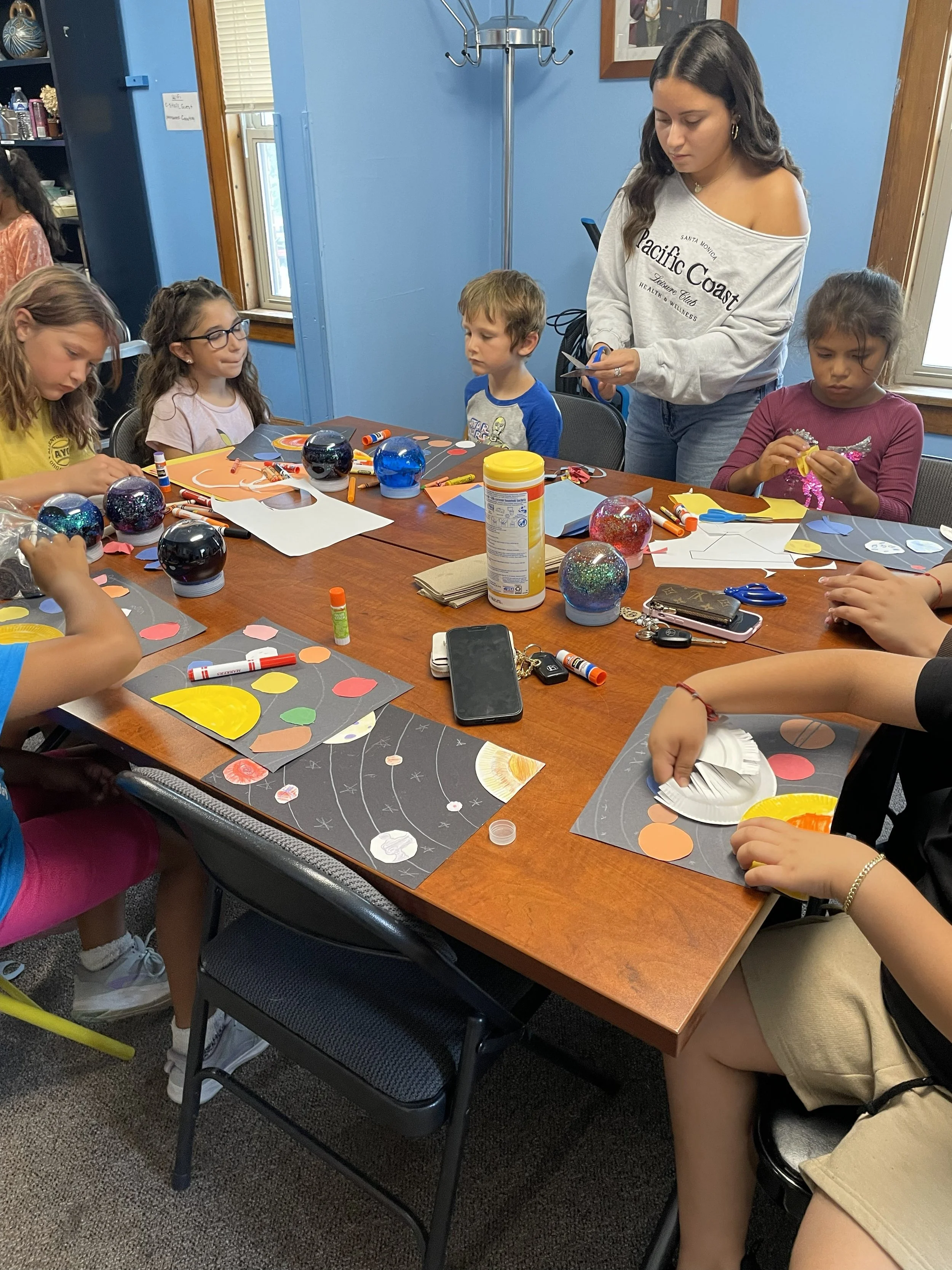 Children and an adult engaged in a craft activity around a wooden table, with colorful paper, glue, scissors, and decorative ornaments on the table.