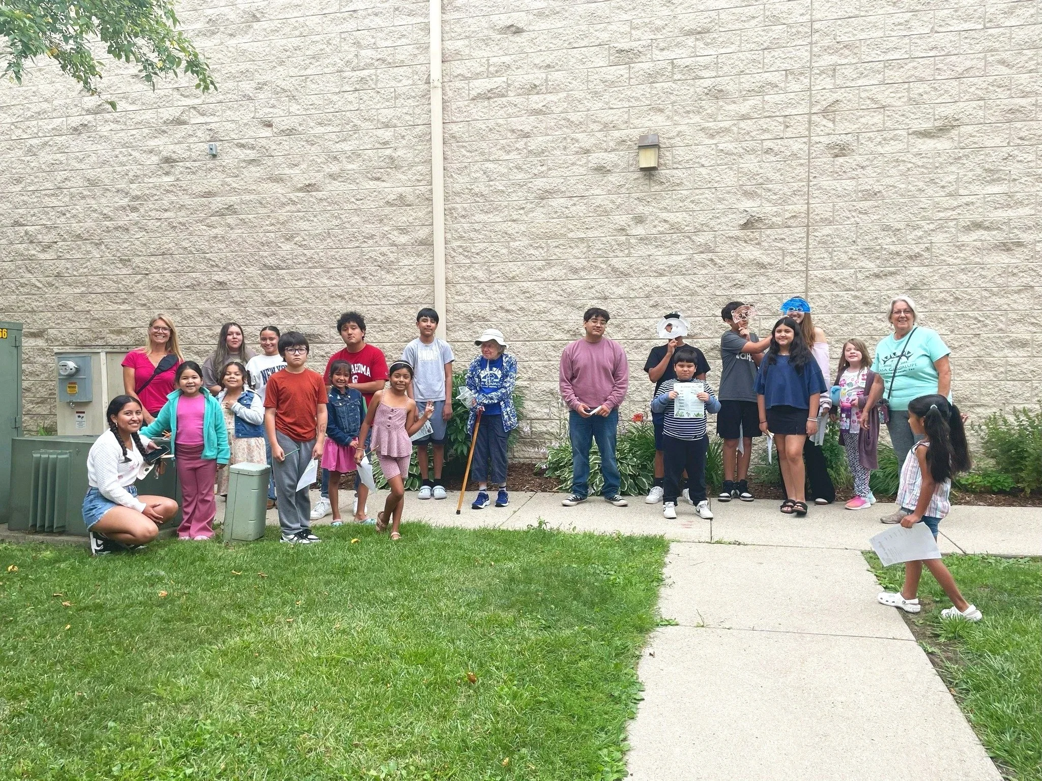 Group of children and adults gathered outside in front of a beige brick wall, most holding sheets of paper, on a sunny day.